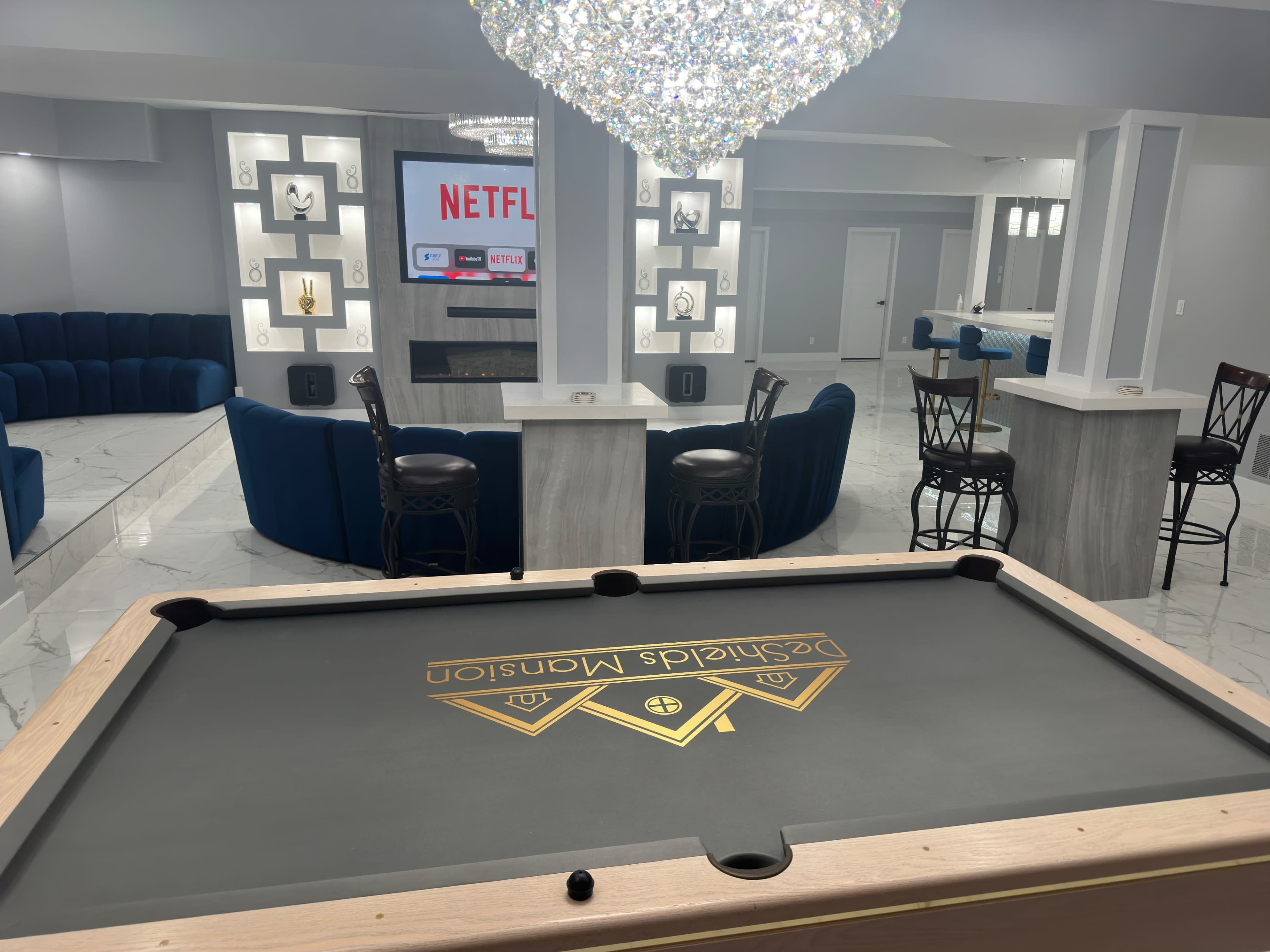 A well-decorated game room featuring a pool table in the foreground, a large television displaying Netflix in the center, and a plush blue seating area in the background.