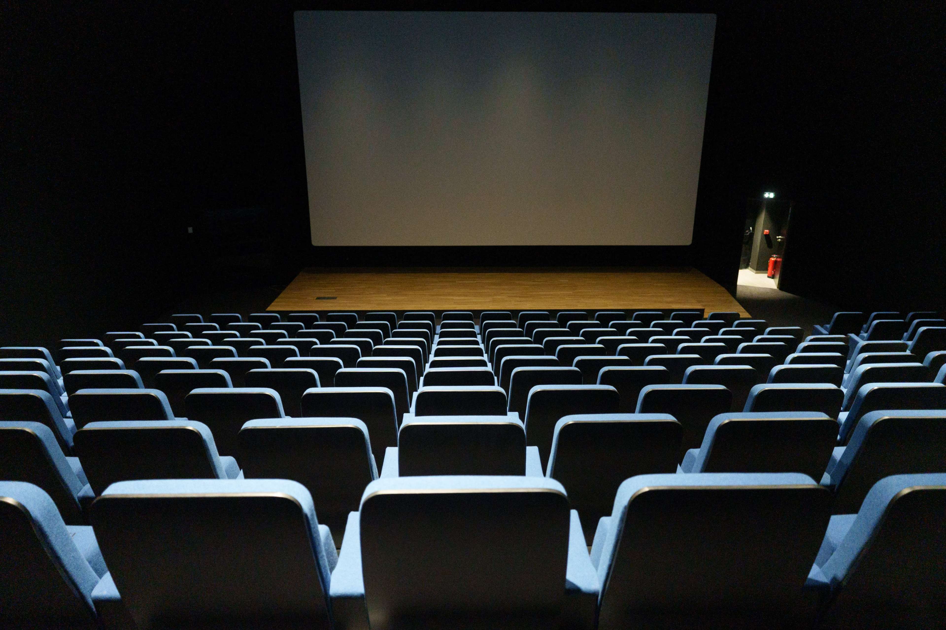 A row of empty blue cinema seats facing a large blank screen in a dark auditorium.