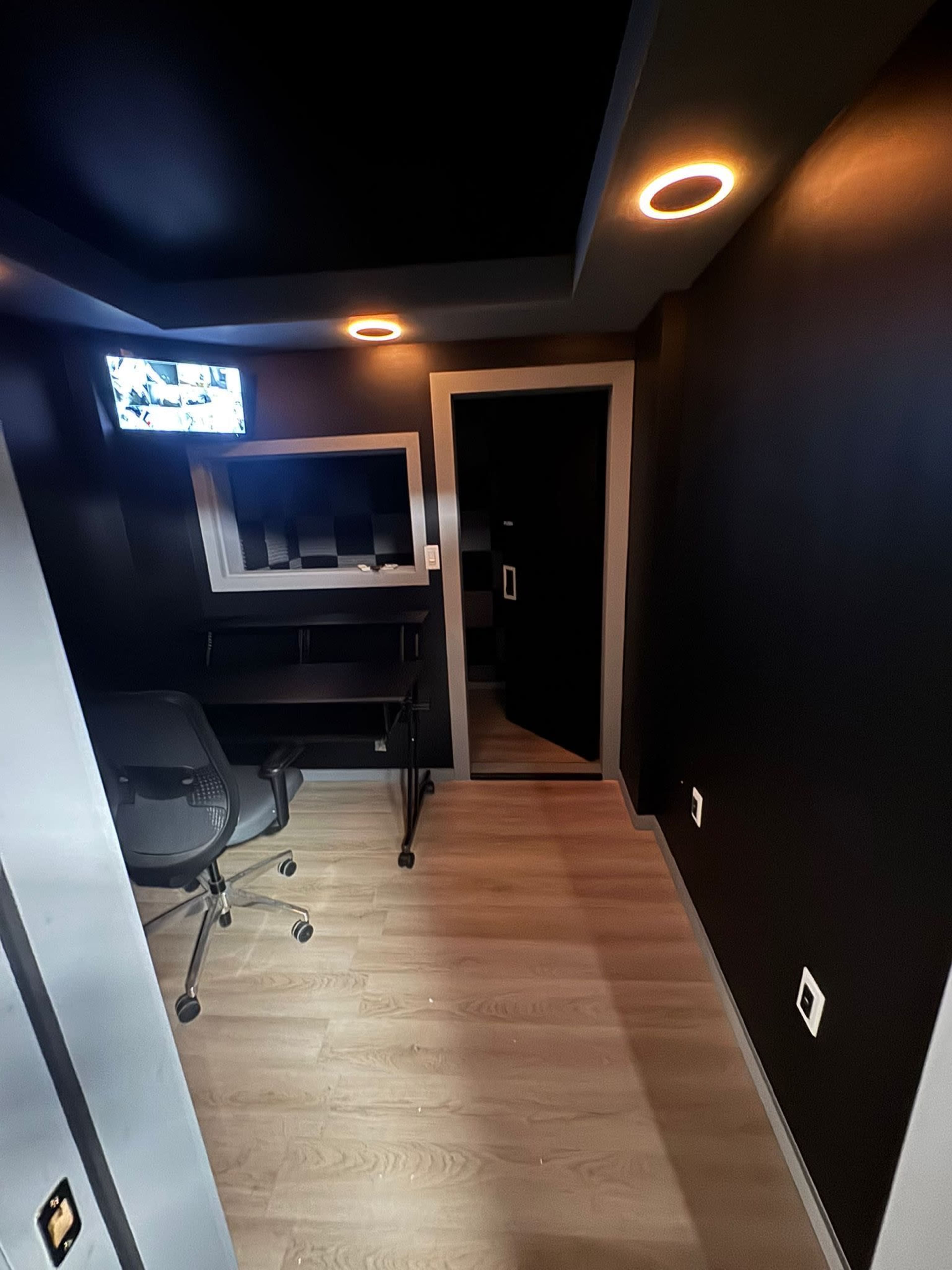 The image shows a small, dark-colored room with a desk, swivel chair, a wall-mounted screen, and a door leading to another space.