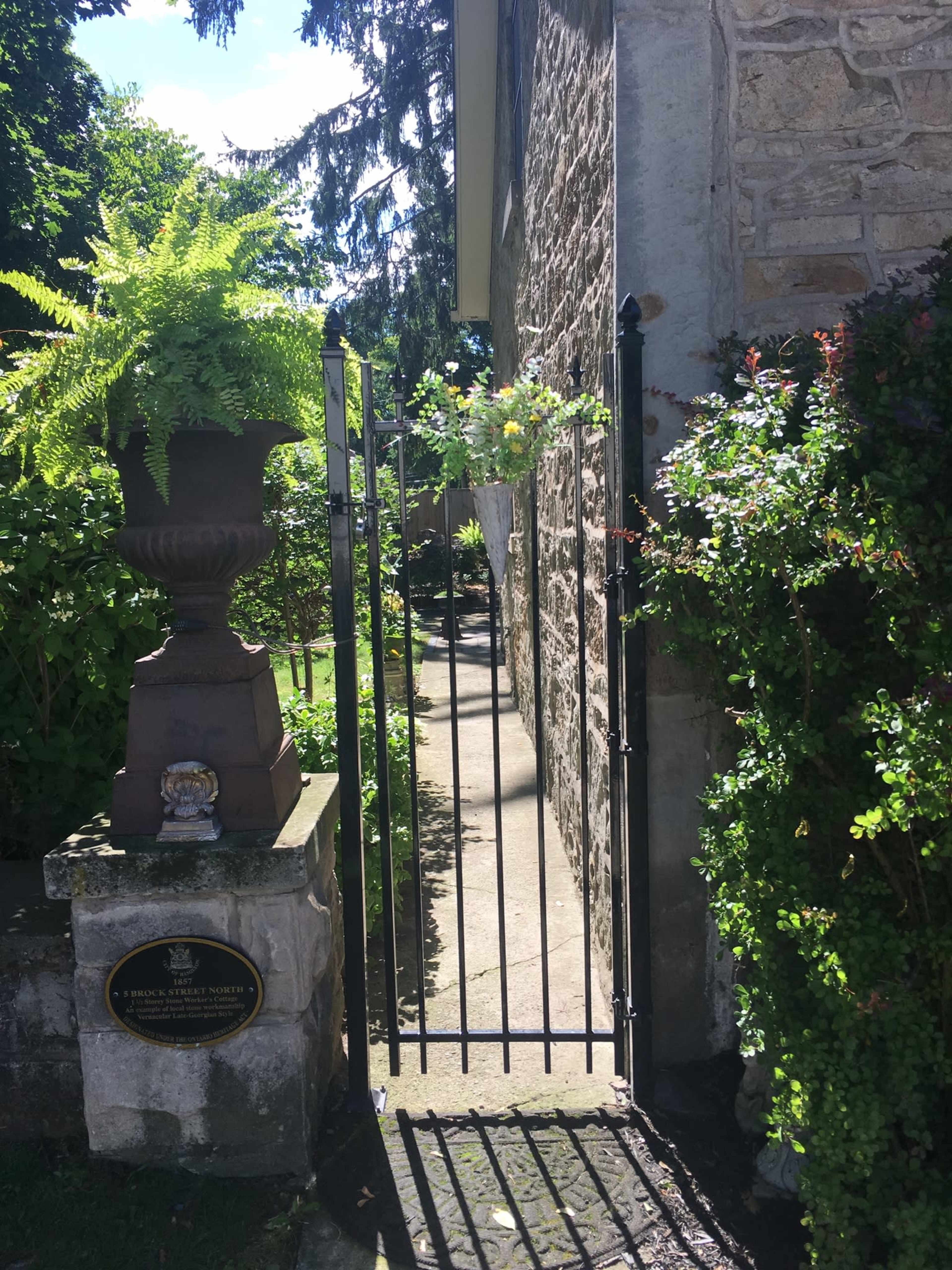 A narrow path leads through an iron gate, flanked by a decorative planter and overgrown greenery beside a stone building.
