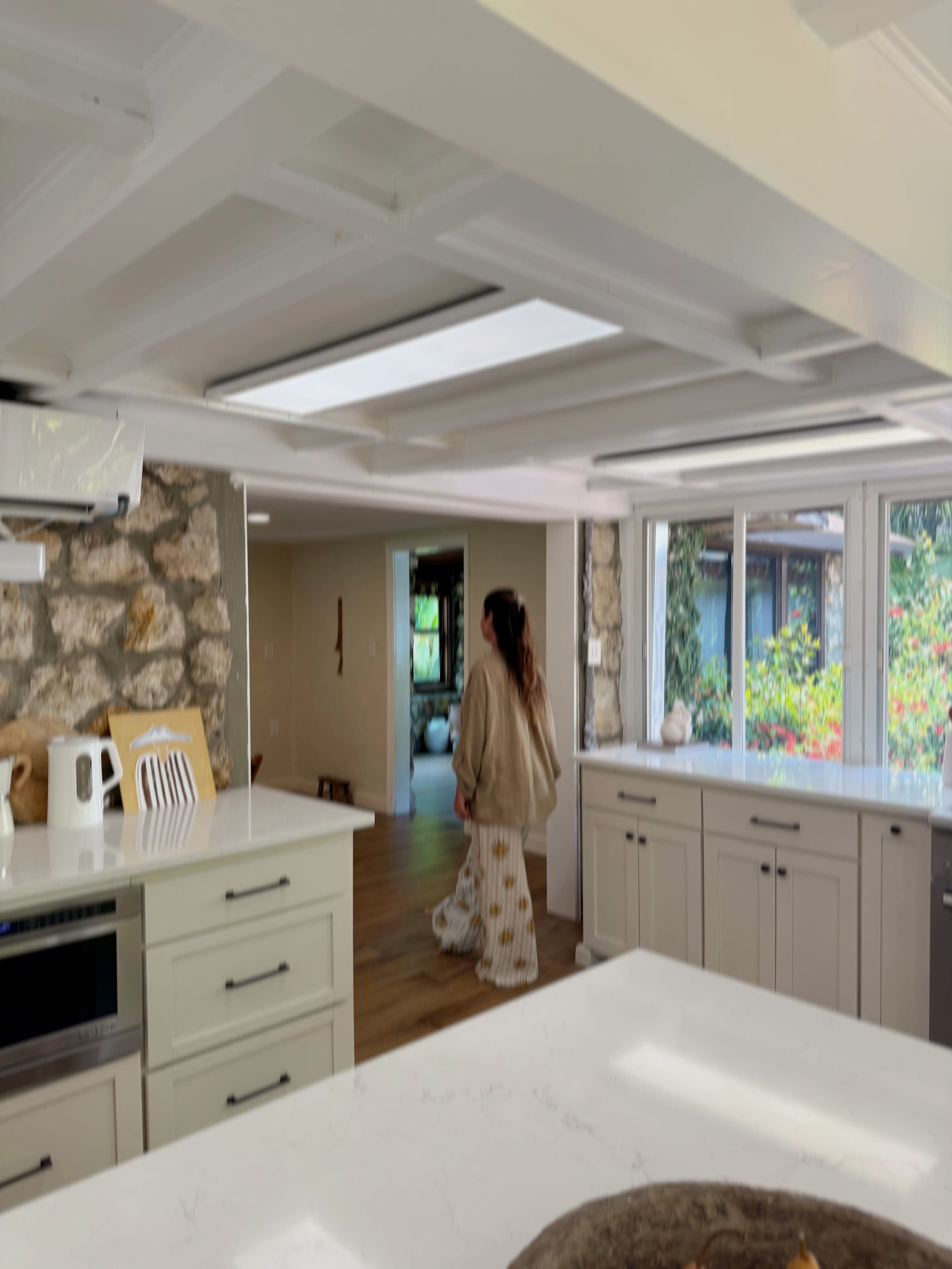 A woman in a long beige robe is walking through a modern kitchen with large windows and wooden flooring.
