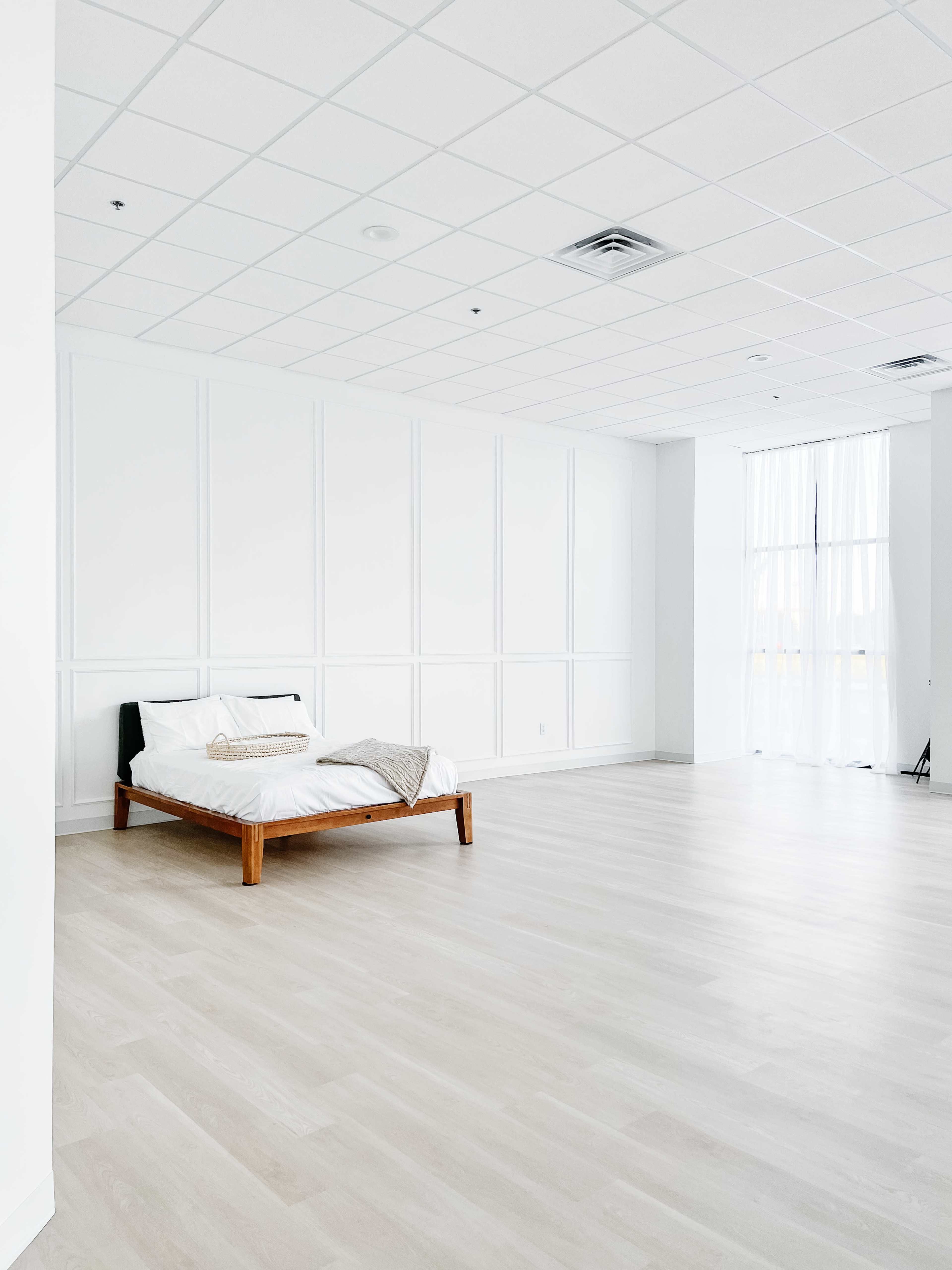 A spacious, minimalistic bedroom with a wooden bed frame positioned against a plain white wall.