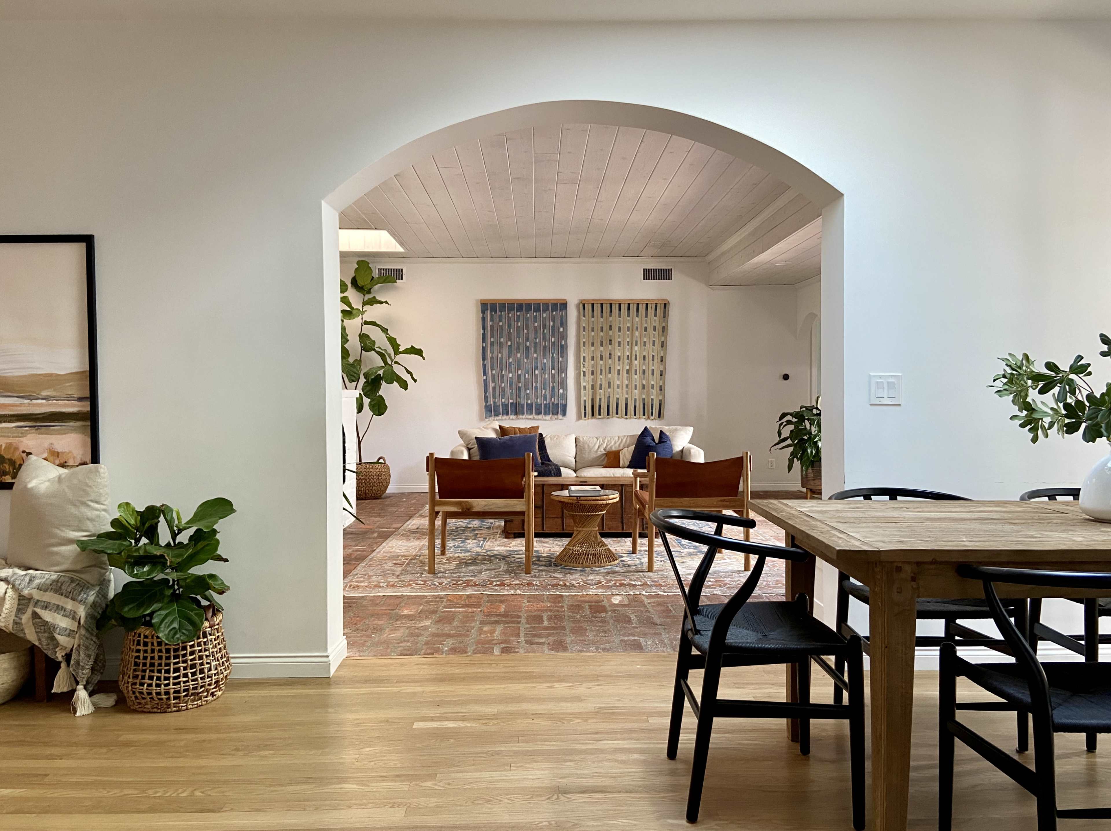 A bright, open living space with an archway leading from a dining area to a cozy seating area featuring a sofa, decorative plants, and artwork on the walls.