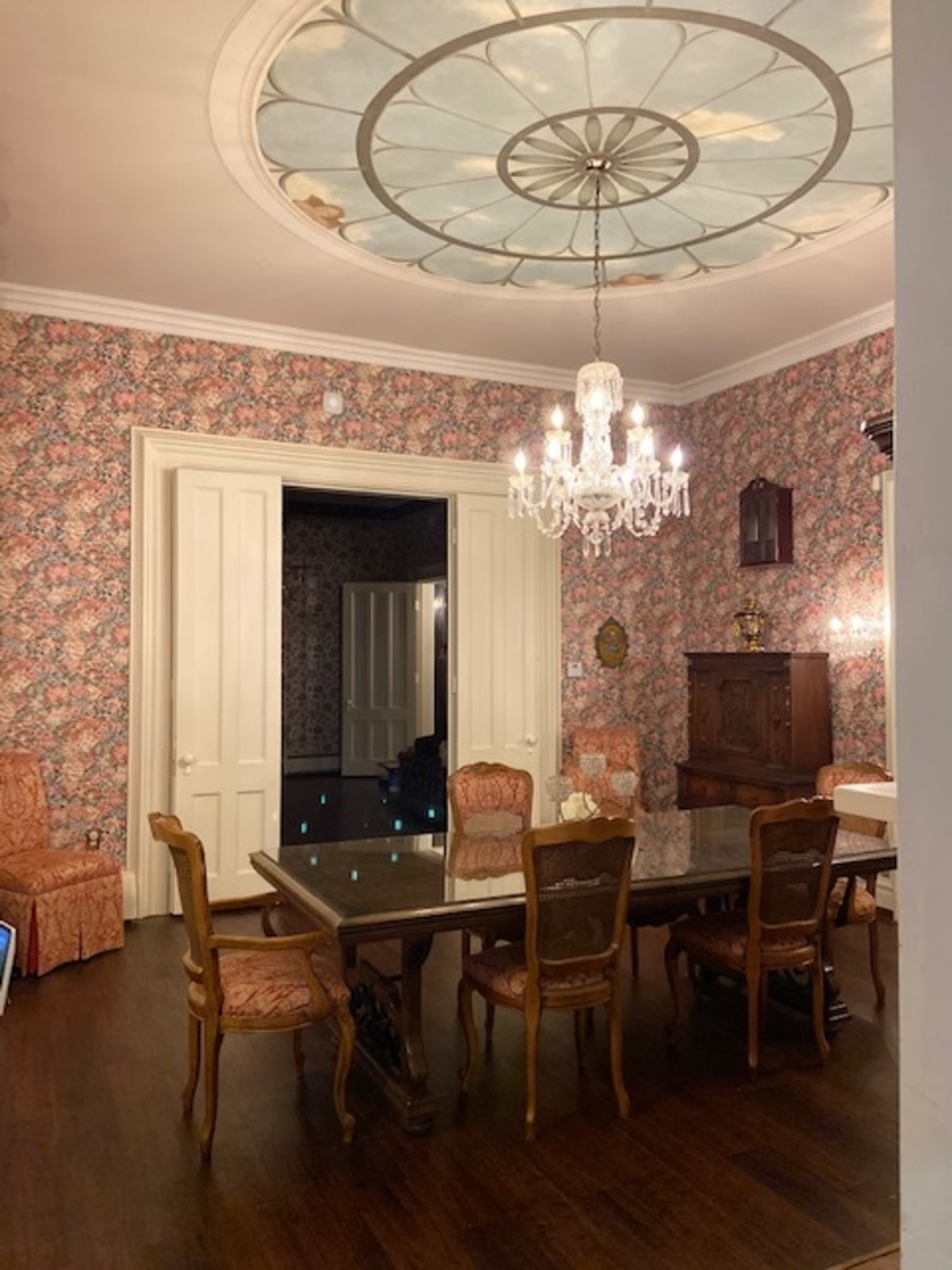 A dining room features a large table surrounded by ornate chairs, with floral wallpaper and an elaborate chandelier overhead.