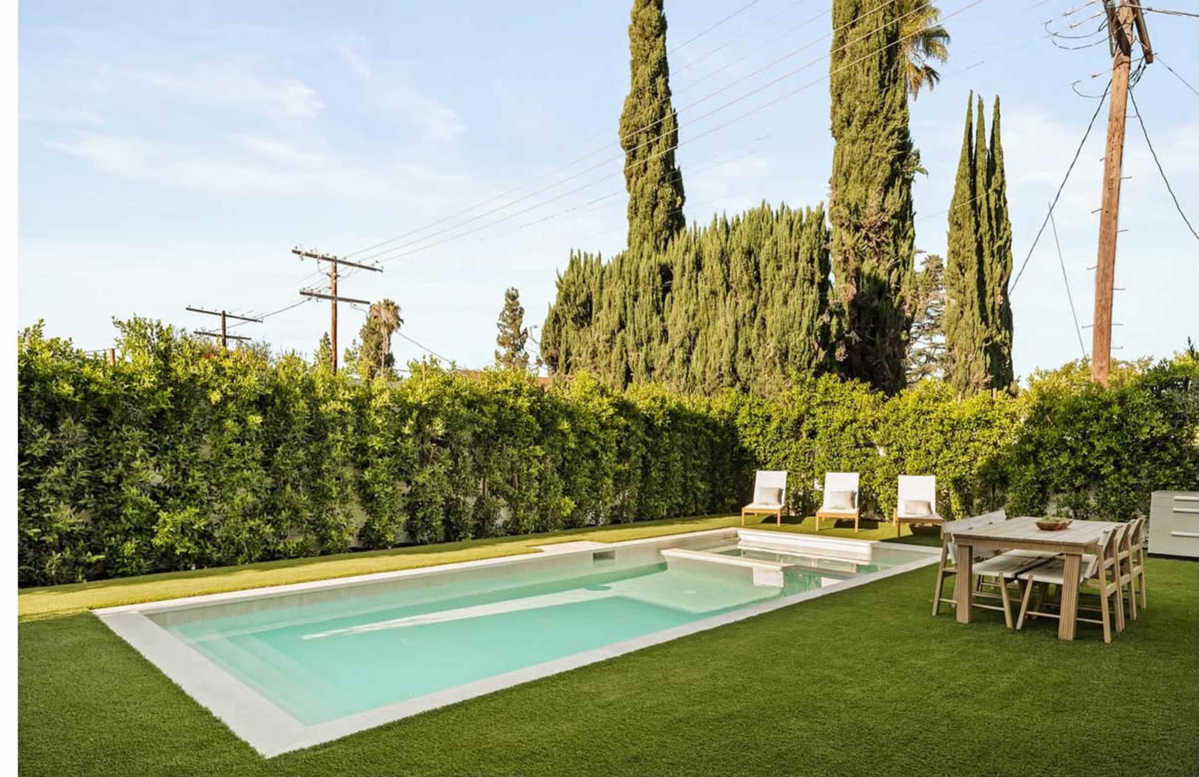 The image shows a backyard featuring a rectangular swimming pool, sun loungers, a dining table, and neatly trimmed hedges against a clear sky.