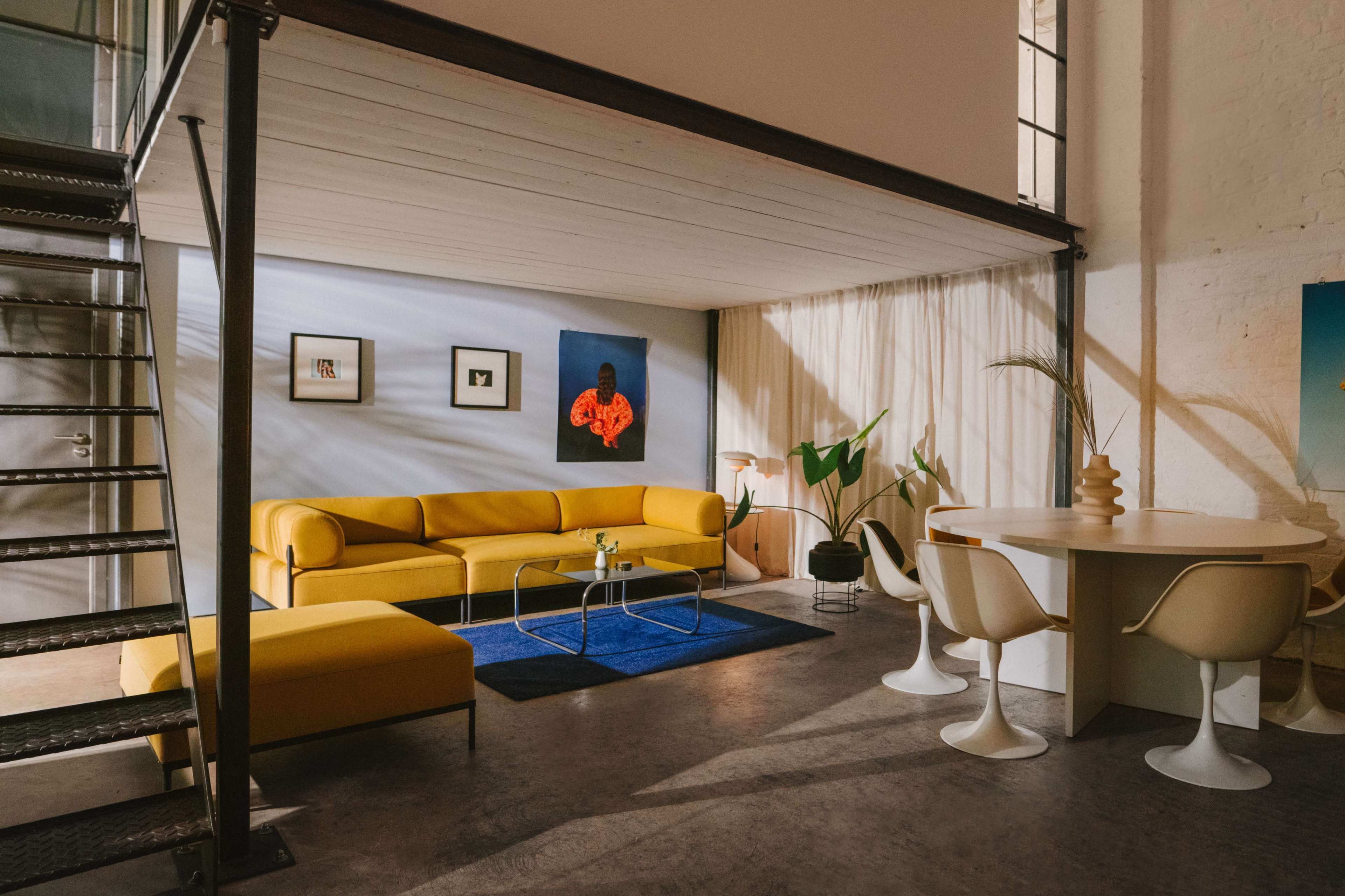 A modern living space featuring a yellow sofa, a round dining table with white chairs, and a staircase leading to an upper level.