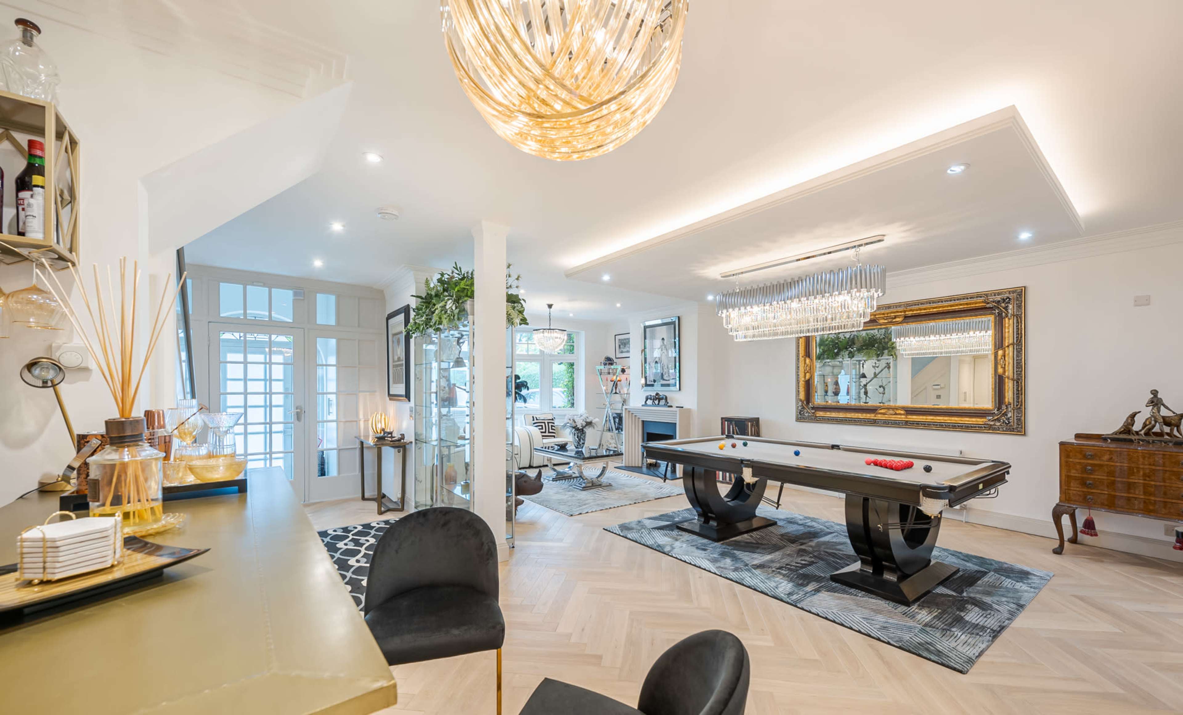 The image displays a modern interior featuring a polished pool table in a spacious living area, illuminated by a large chandelier and accented with stylish decor.