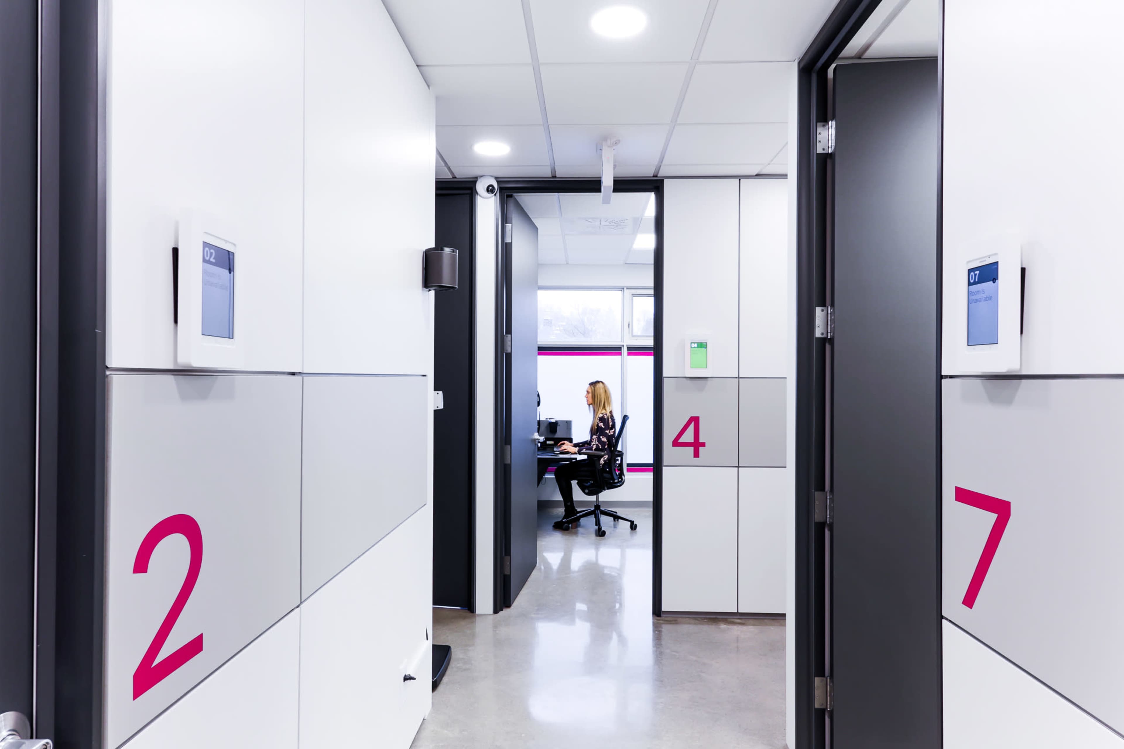 The image shows a modern office corridor with room numbers displayed on the walls and a person seated at a desk within one of the visible rooms.