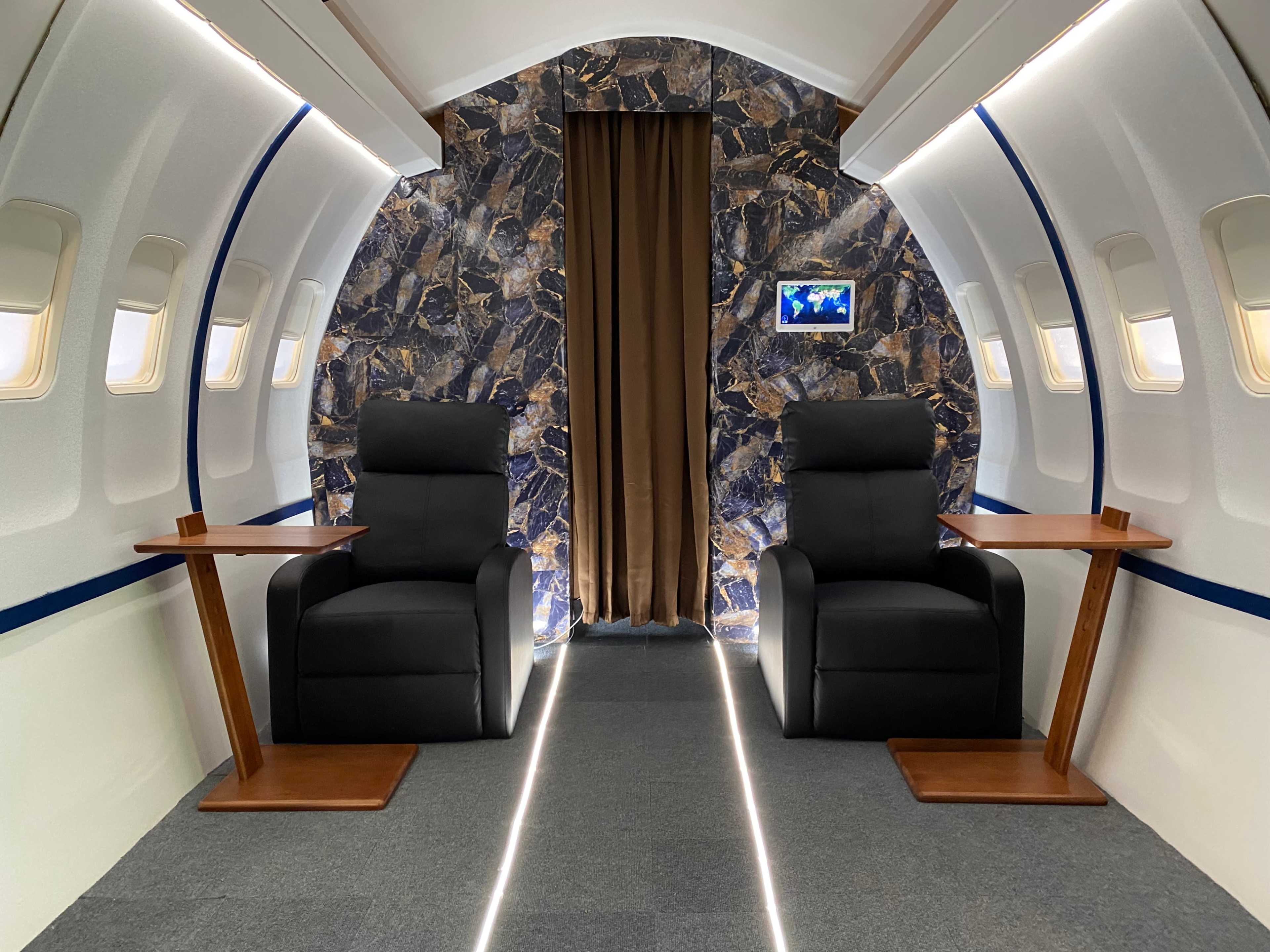 The interior of a private jet showcases two black reclining chairs flanking a central aisle, with a patterned wall and soft lighting.