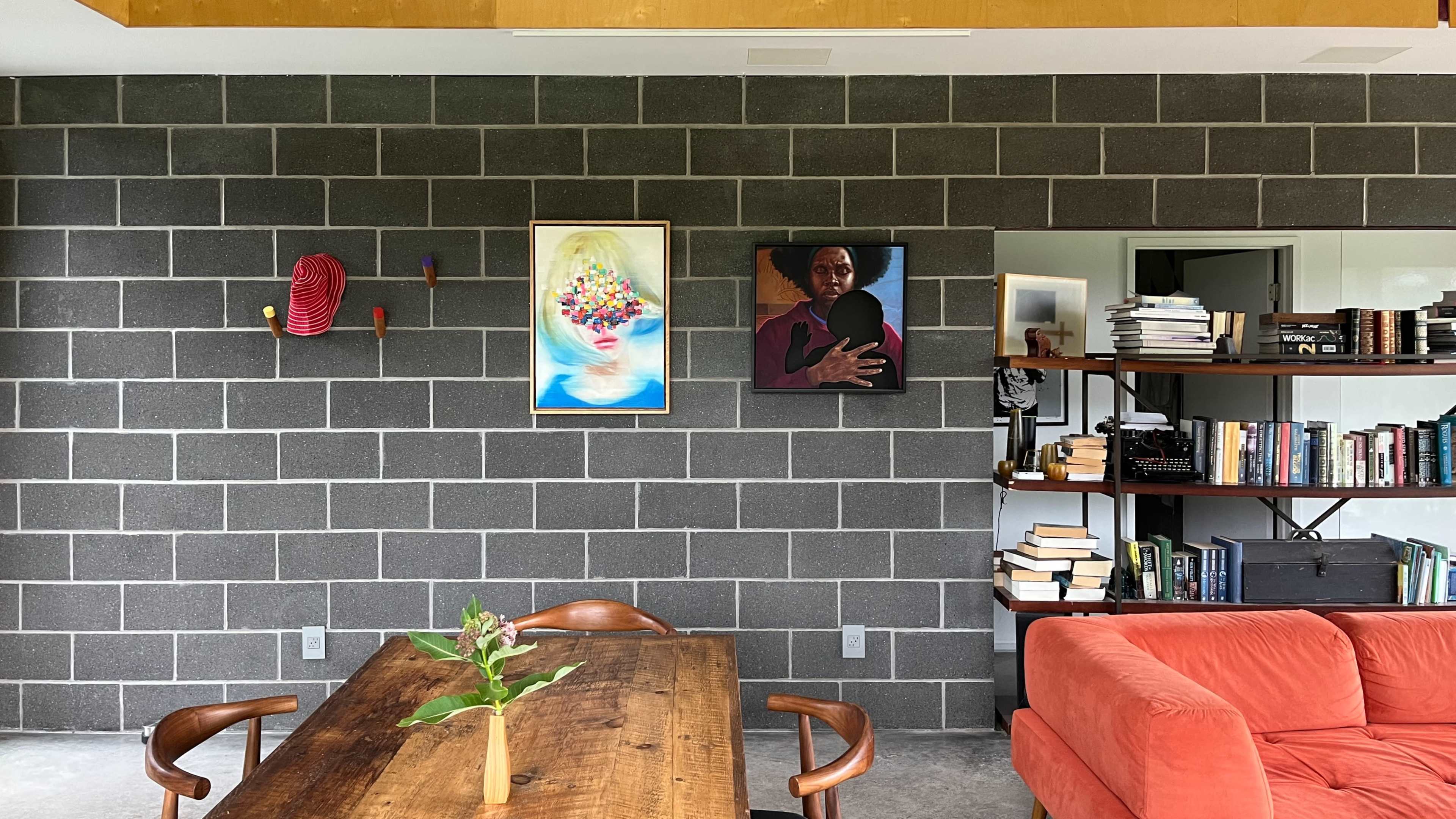A living space with a gray block wall featuring two framed artworks, a wooden dining table with a small plant, and an orange couch beside a bookshelf filled with books.