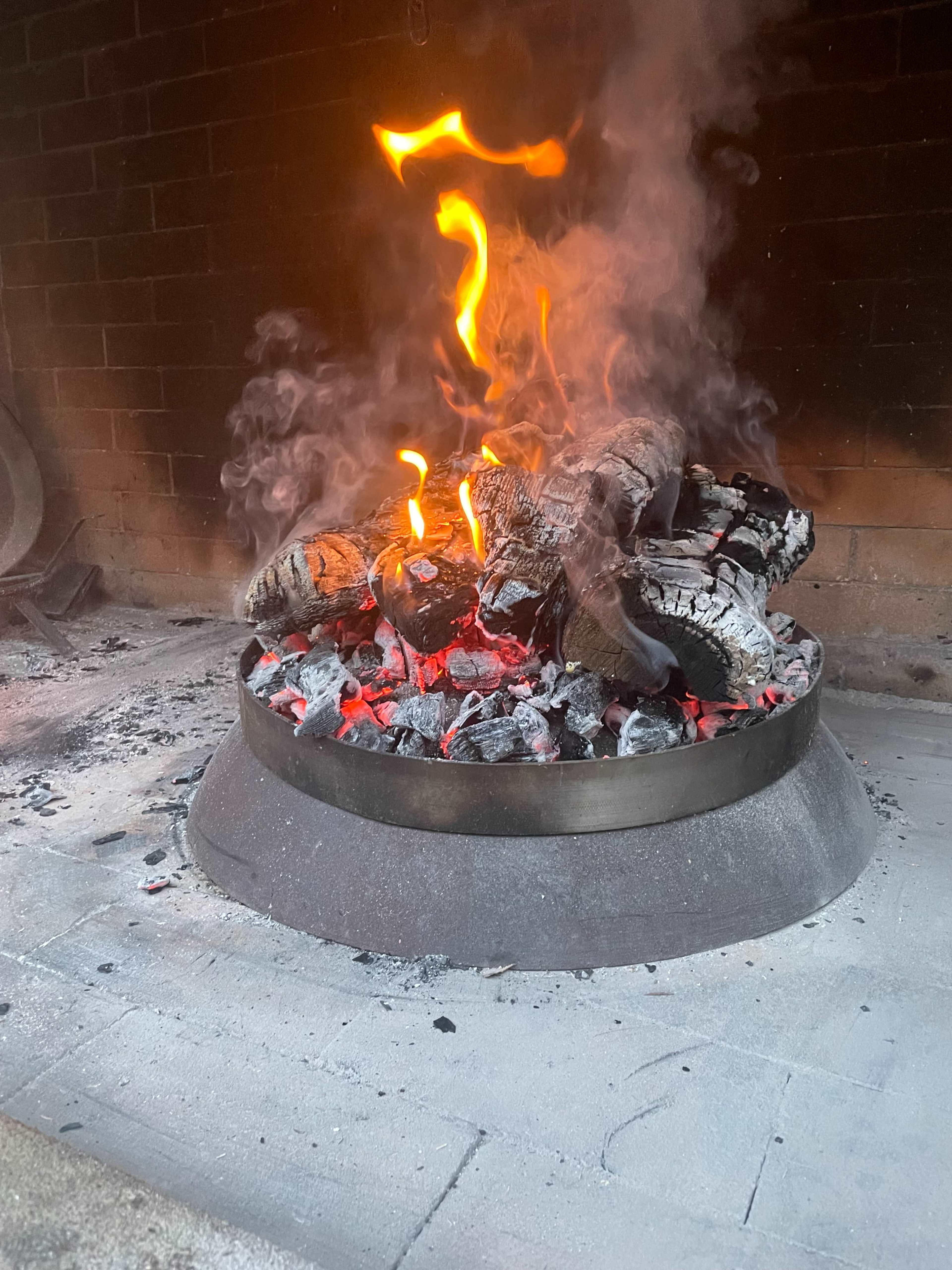 A fire burns brightly in a circular metal pit, surrounded by glowing embers and smoke.