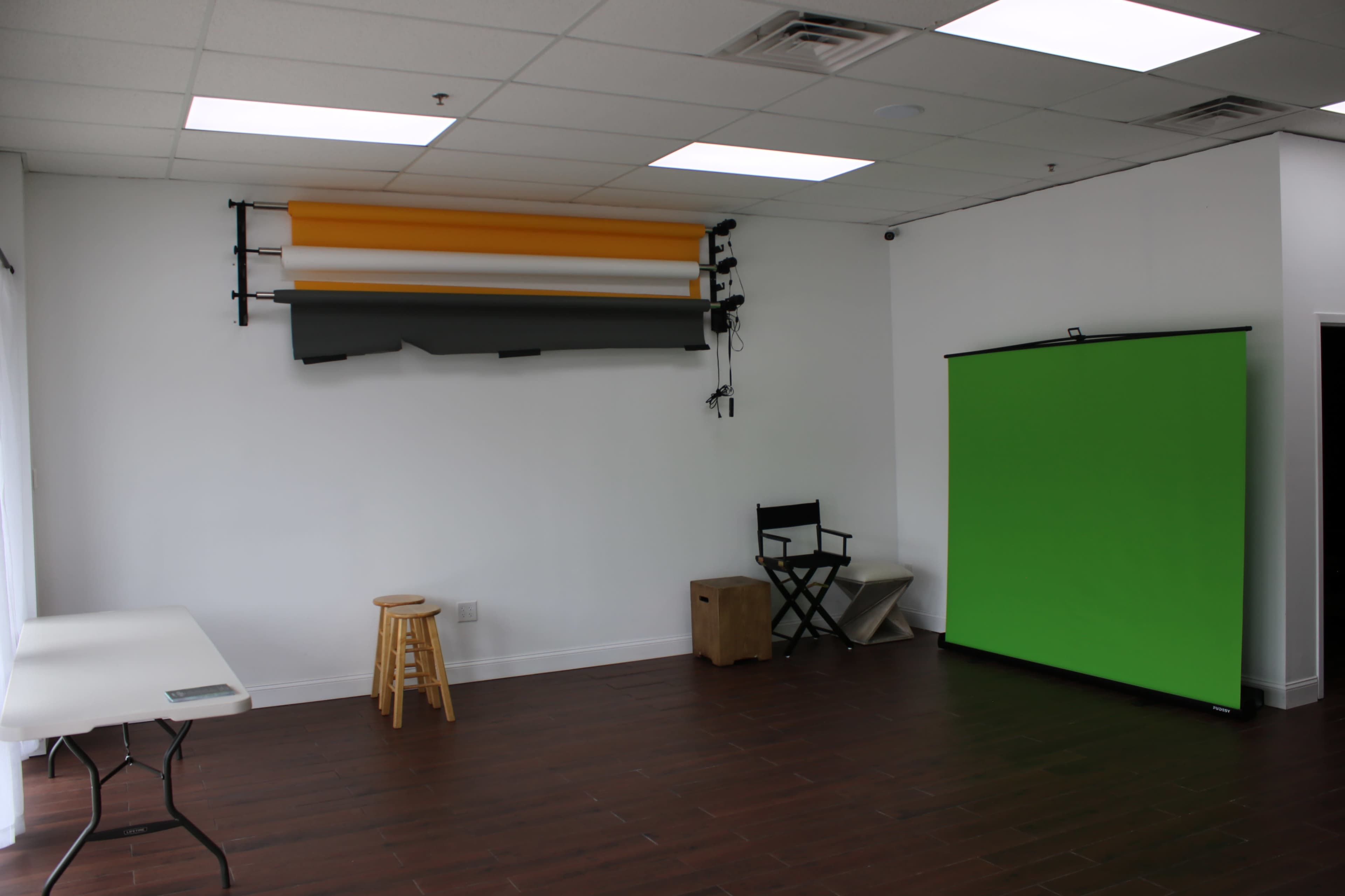 A bright studio space with a green backdrop on one side, several rolls of colored backdrops mounted on the wall, a stool, and a folding table.