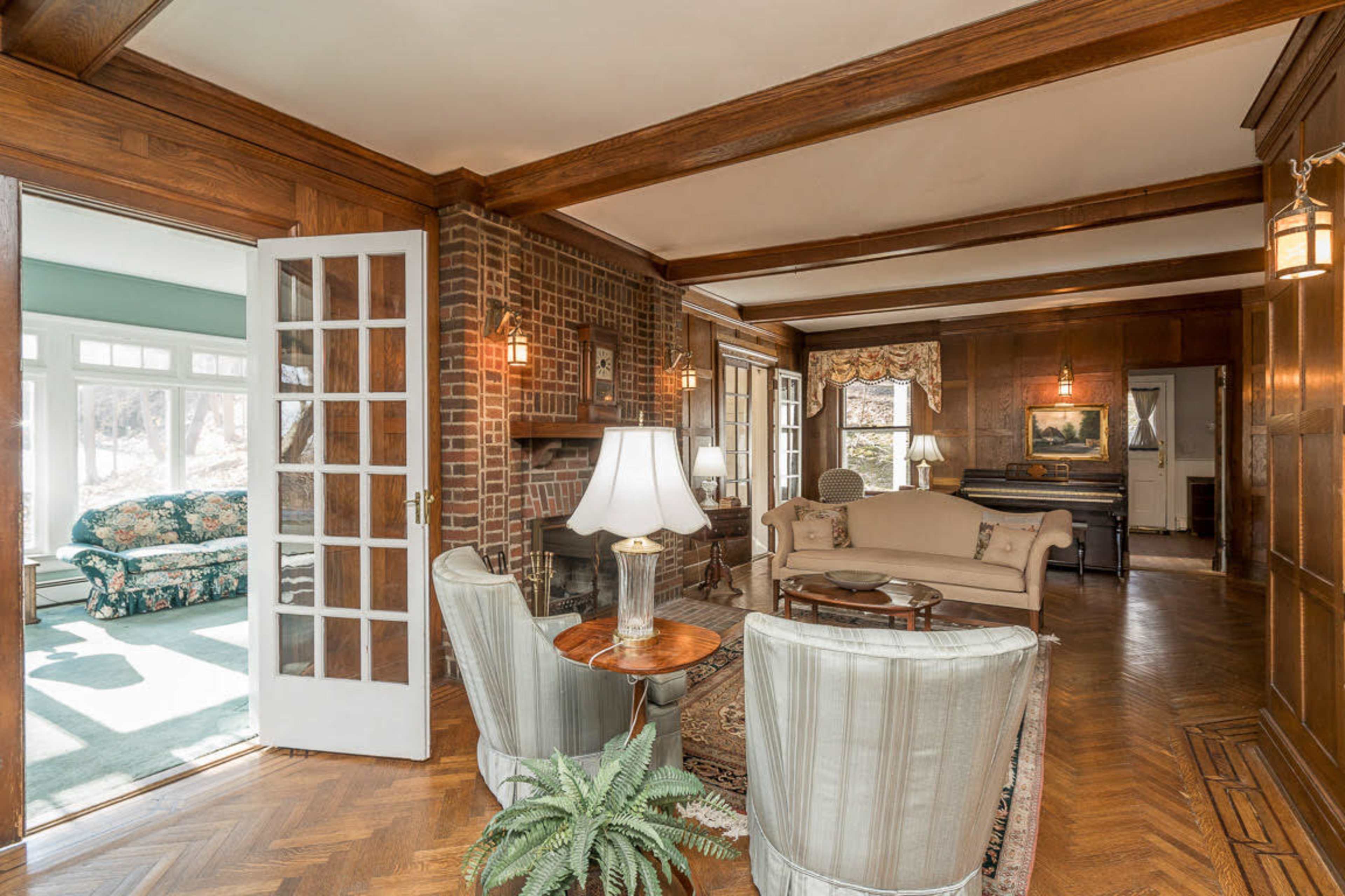The image shows a living room with wooden beams, a brick fireplace, and comfortable seating along with a view into a sunlit room beyond.