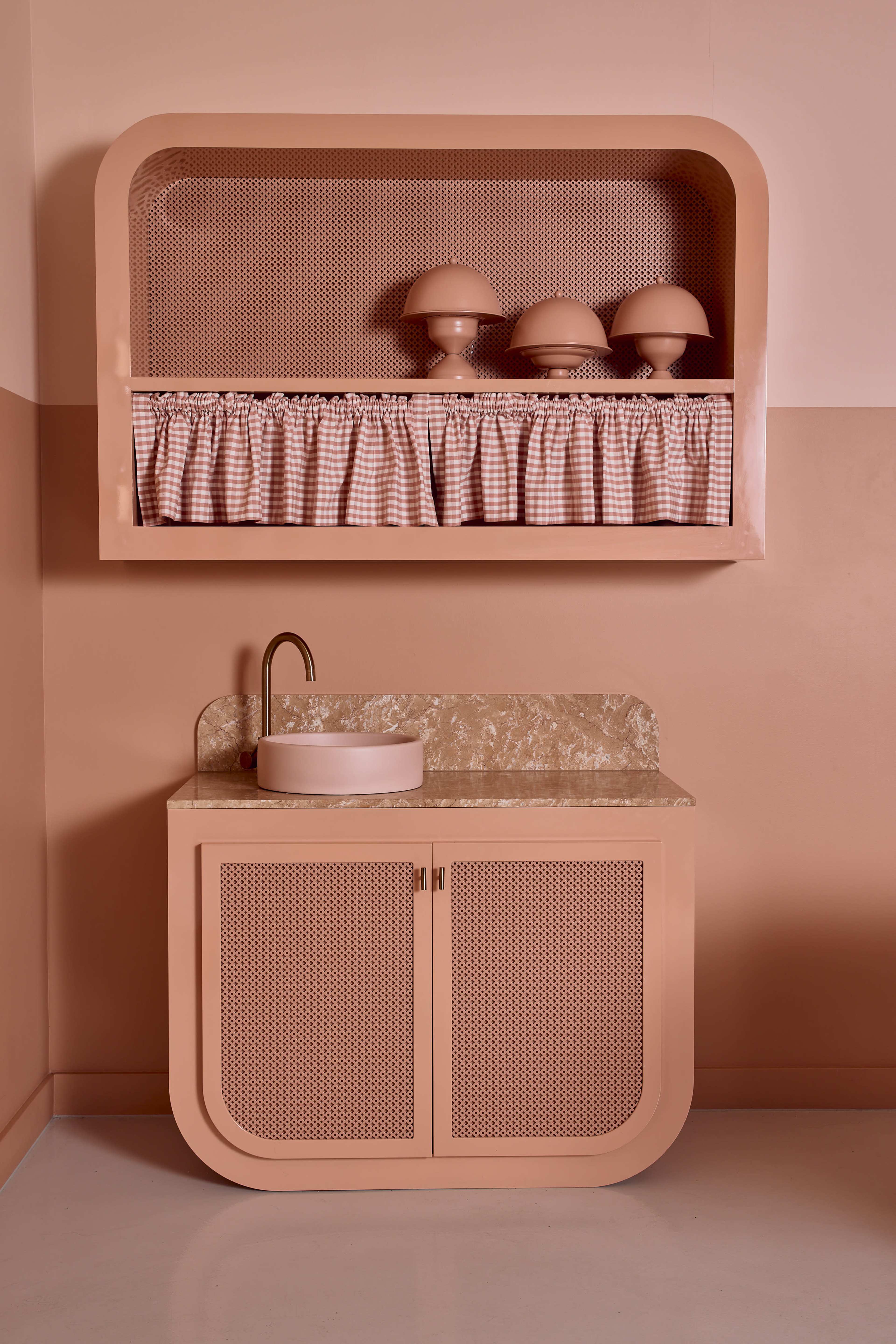 The image shows a minimalist kitchen area featuring a rounded peach-colored cabinet with a marble countertop and a sink, accompanied by a matching shelf above adorned with decorative items.