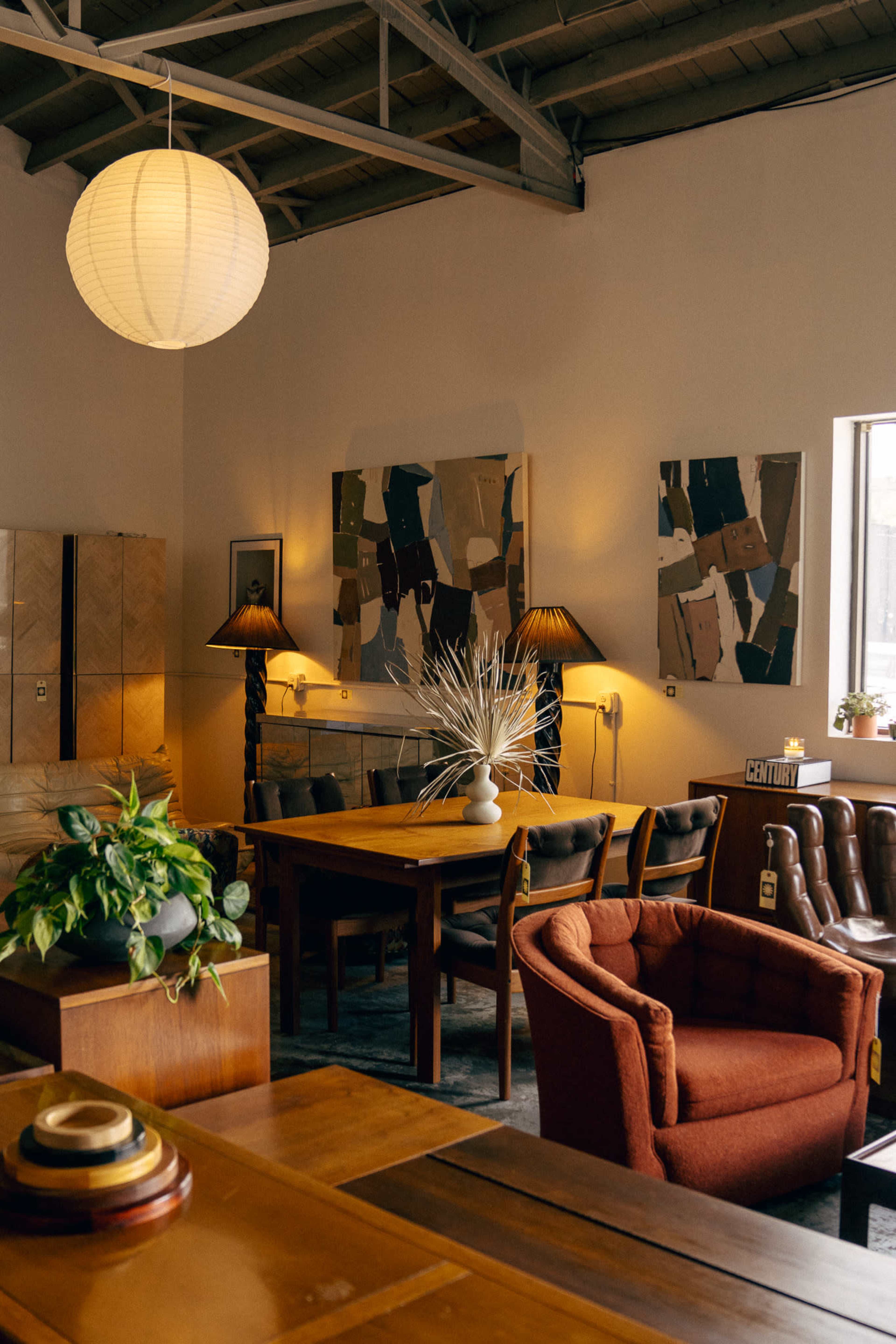 The image shows a stylish interior of a furniture showroom featuring wooden tables, chairs, and wall art, illuminated by warm lighting.