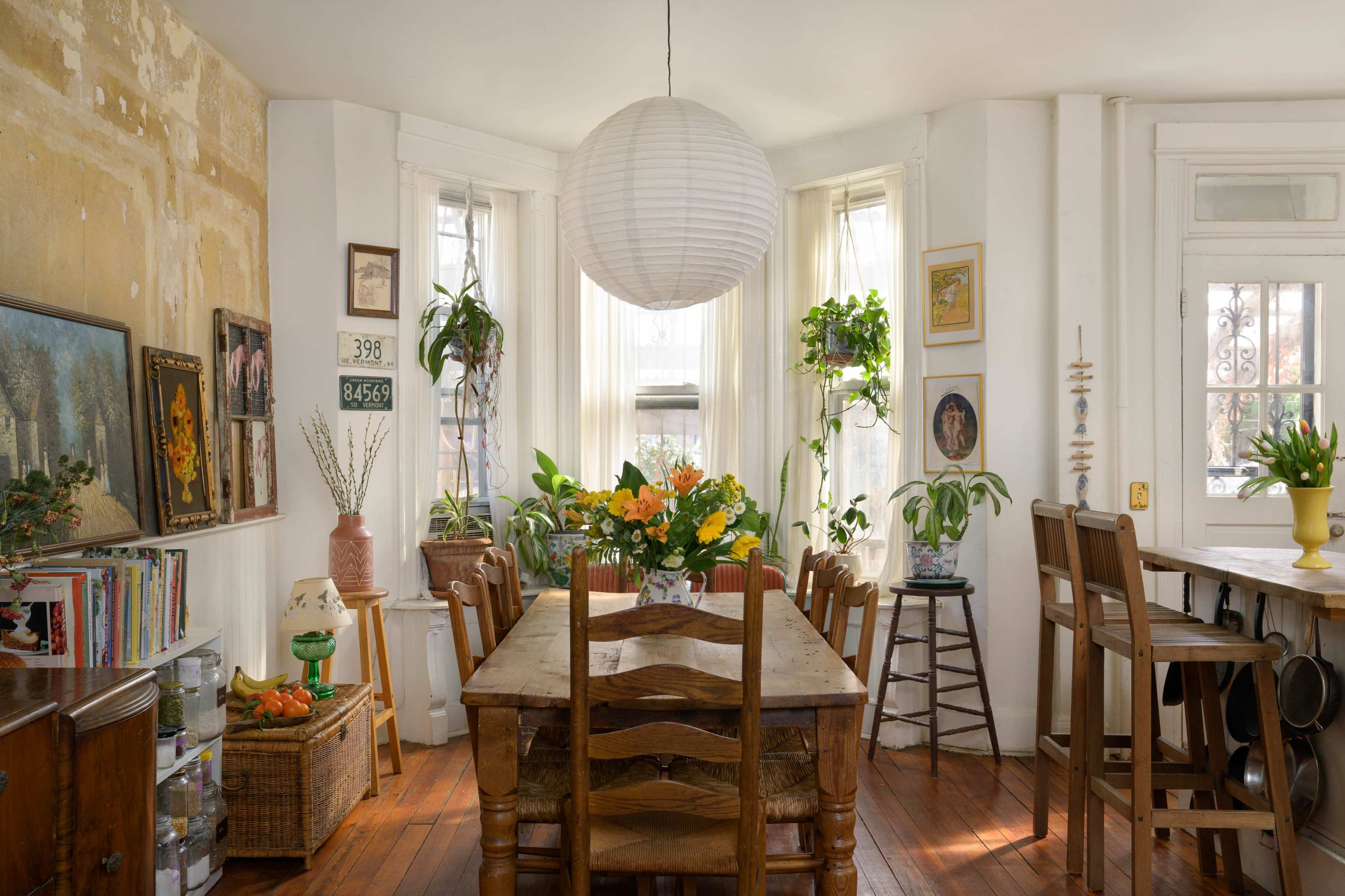 A bright dining area features a large wooden table surrounded by chairs, with potted plants and artwork adorning the walls.
