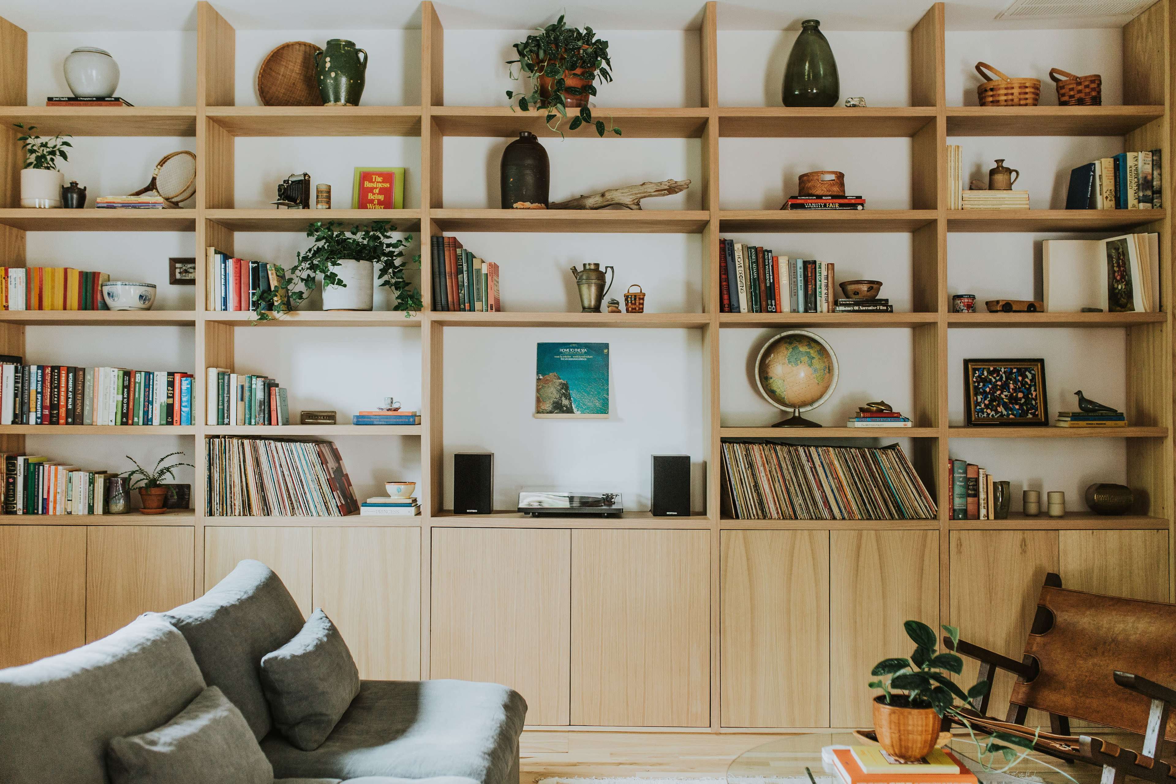 A wooden bookshelf filled with various books, plants, and decor items stands against a wall in a cozy living room, accompanied by a gray sofa and a coffee table.