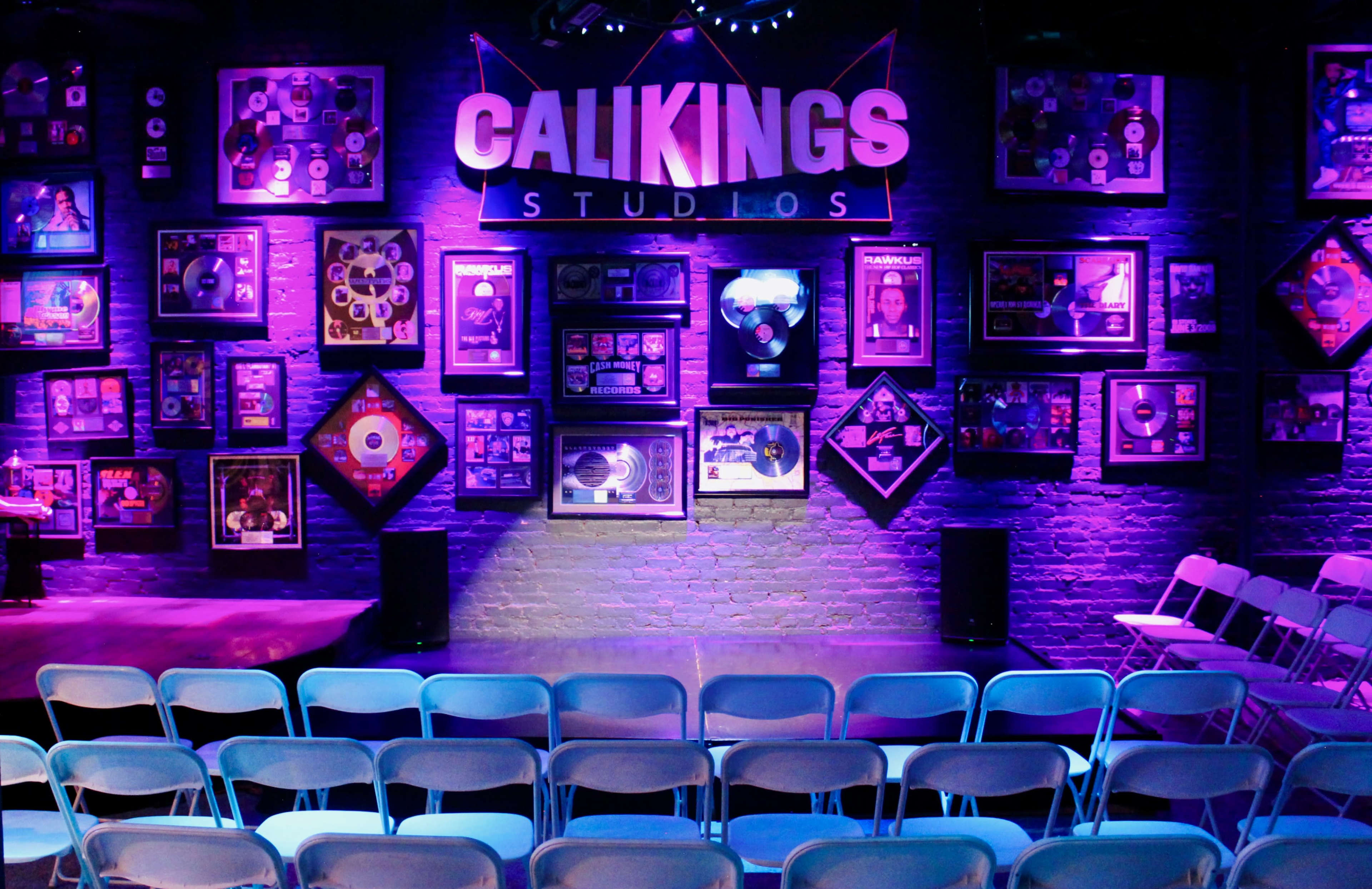 A music studio decorated with framed records and awards on a purple-lit wall, with rows of white chairs arranged in front.
