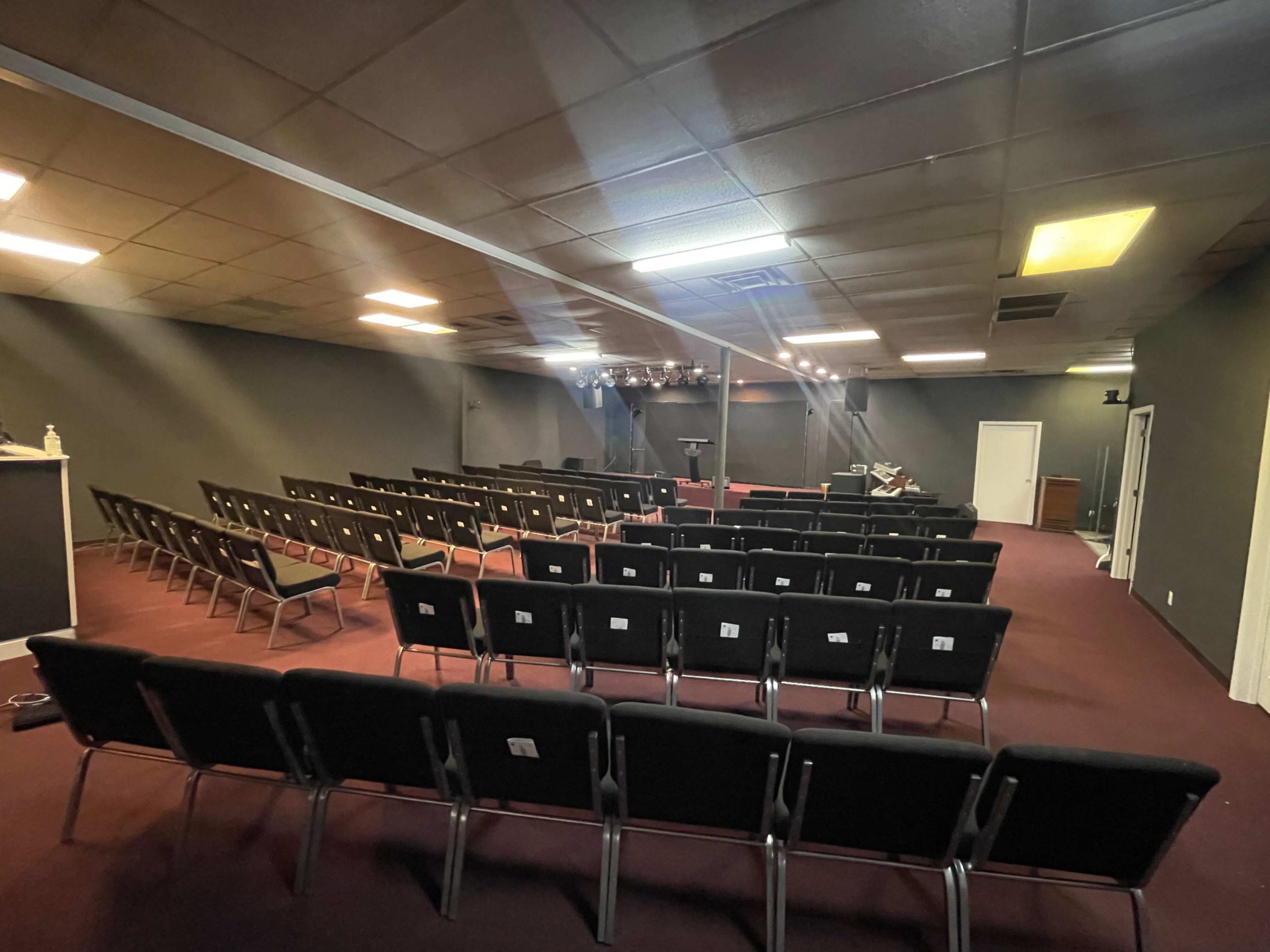 The image shows a sparsely furnished auditorium with rows of black chairs facing a small stage and lighting setup.