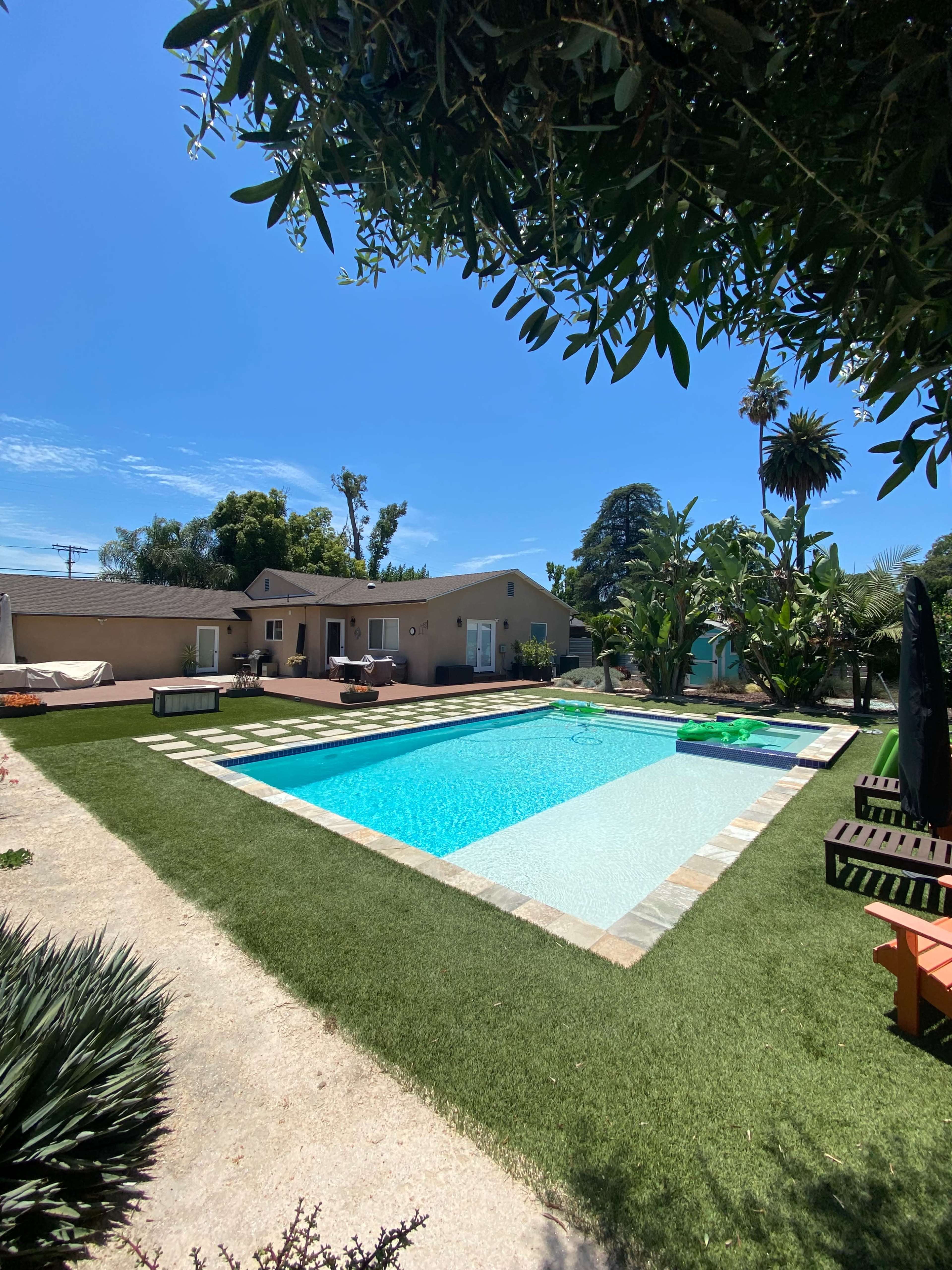 A clear blue swimming pool is surrounded by a landscaped yard featuring grass and palm trees, with a house in the background.
