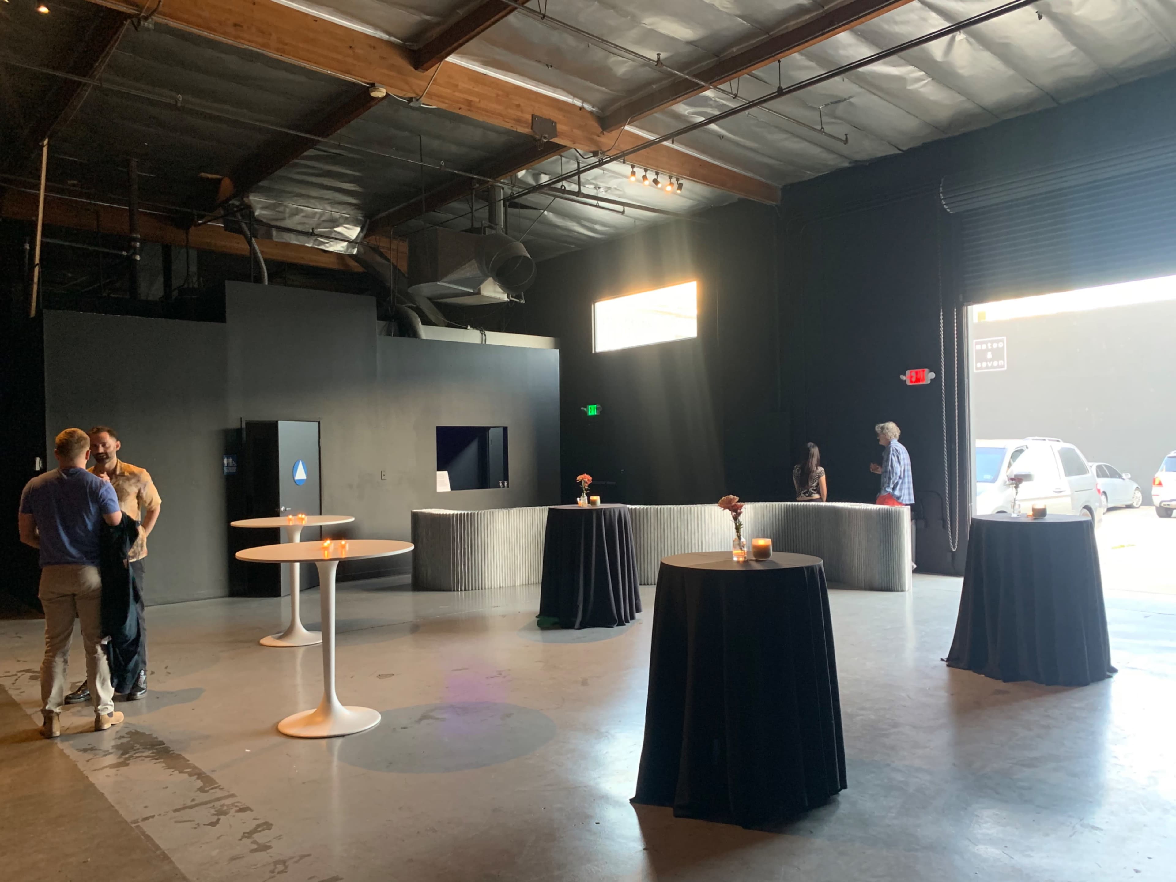 The scene shows a dimly lit event space with several high tables, a check-in area, and a large opening that leads outside.