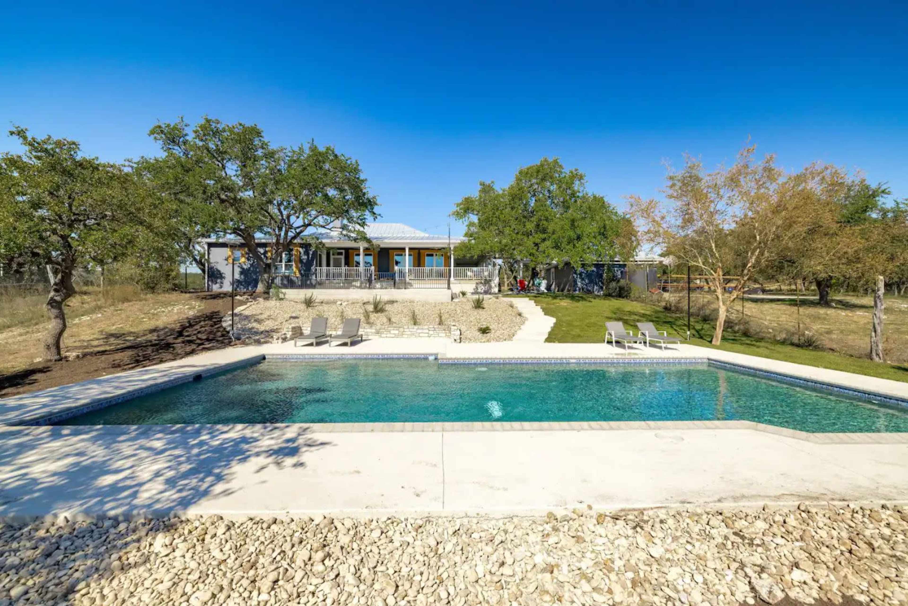 The image shows a swimming pool surrounded by rocky landscaping and a grassy area, with a house in the background flanked by trees.