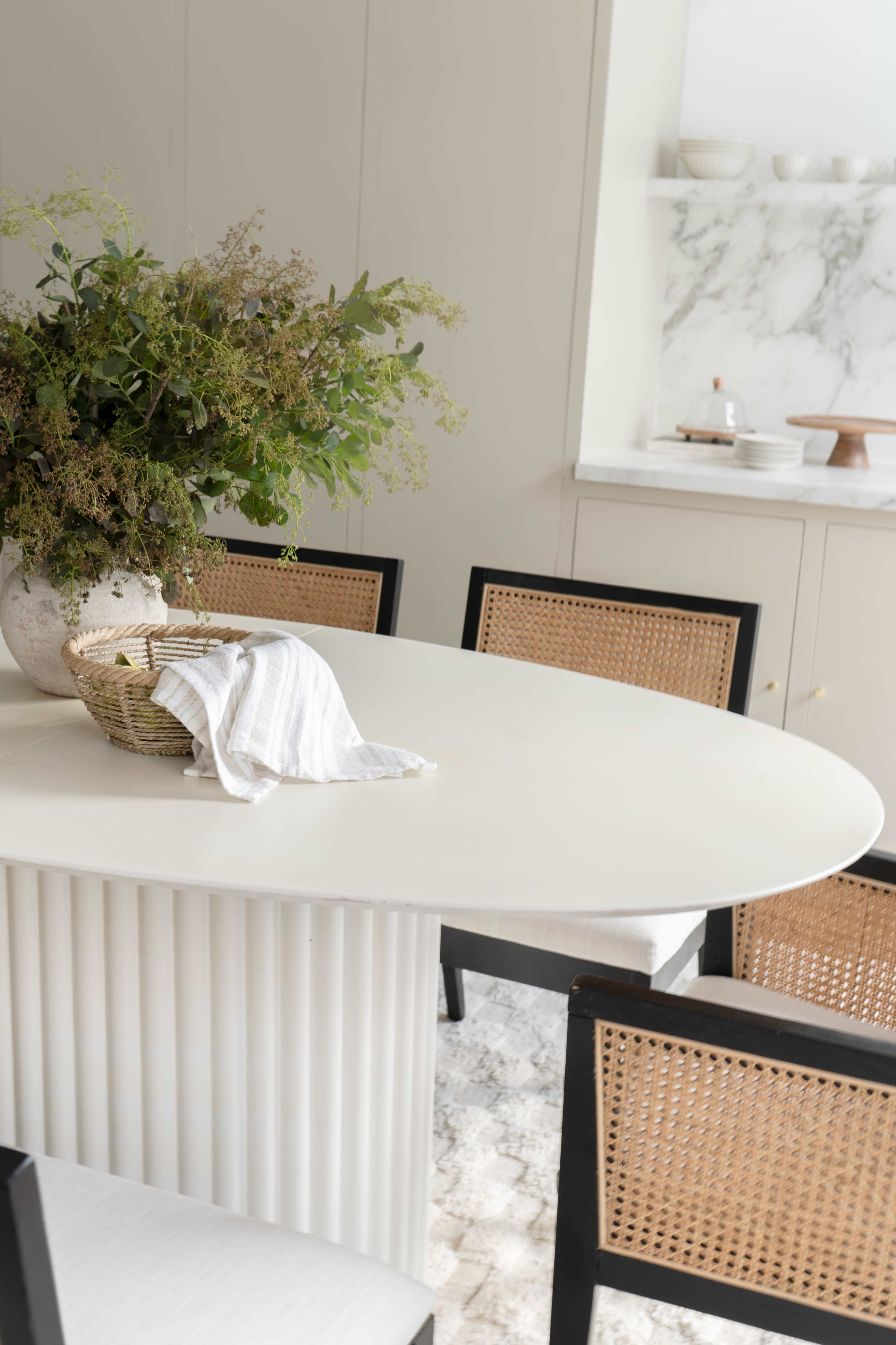 A round, white dining table is set with a vase of greenery and a woven basket, surrounded by four rattan-backed chairs in a light-filled room.