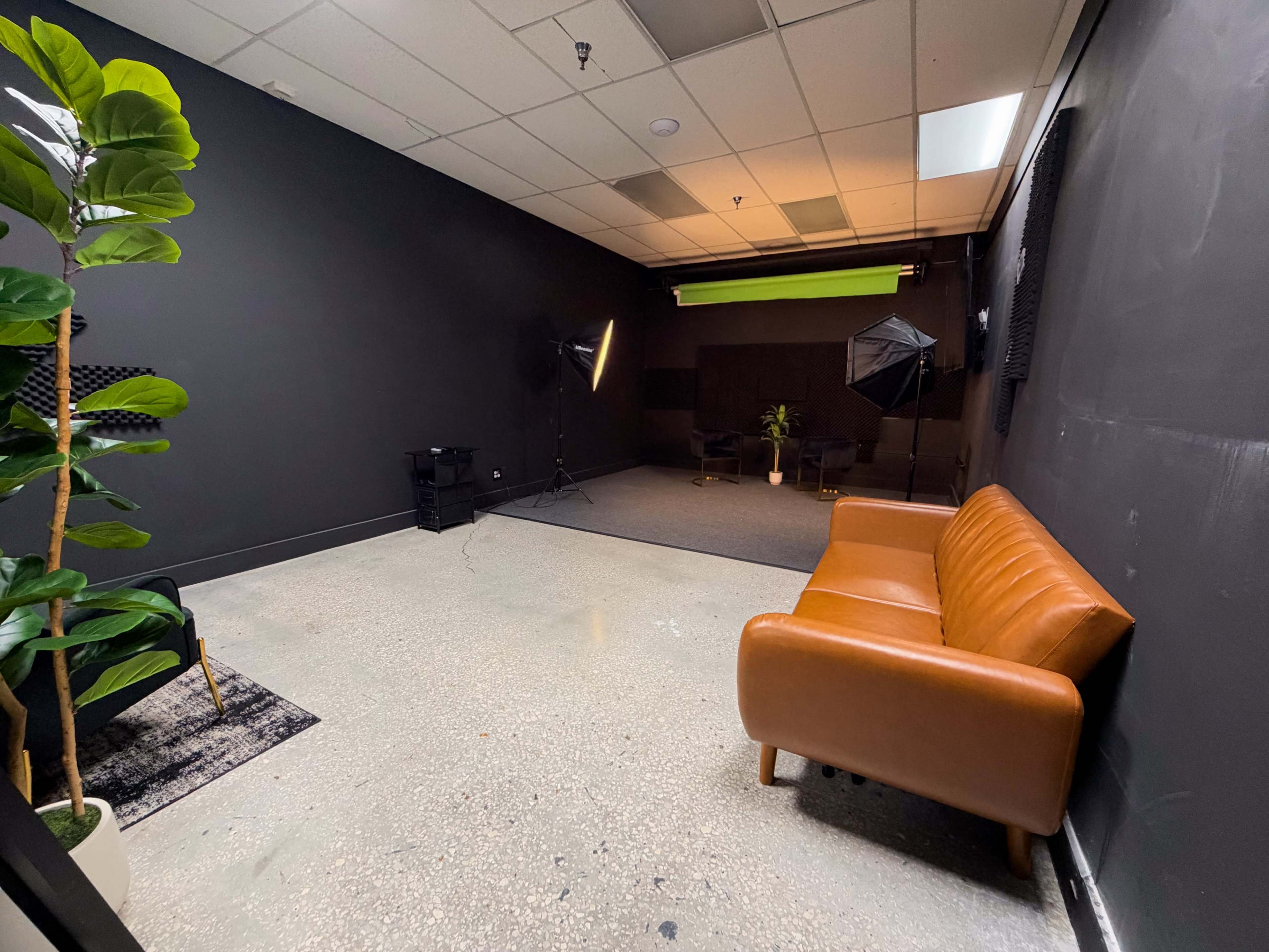 The image depicts a sparsely furnished photo studio featuring a brown leather couch, potted plant, and professional lighting equipment.