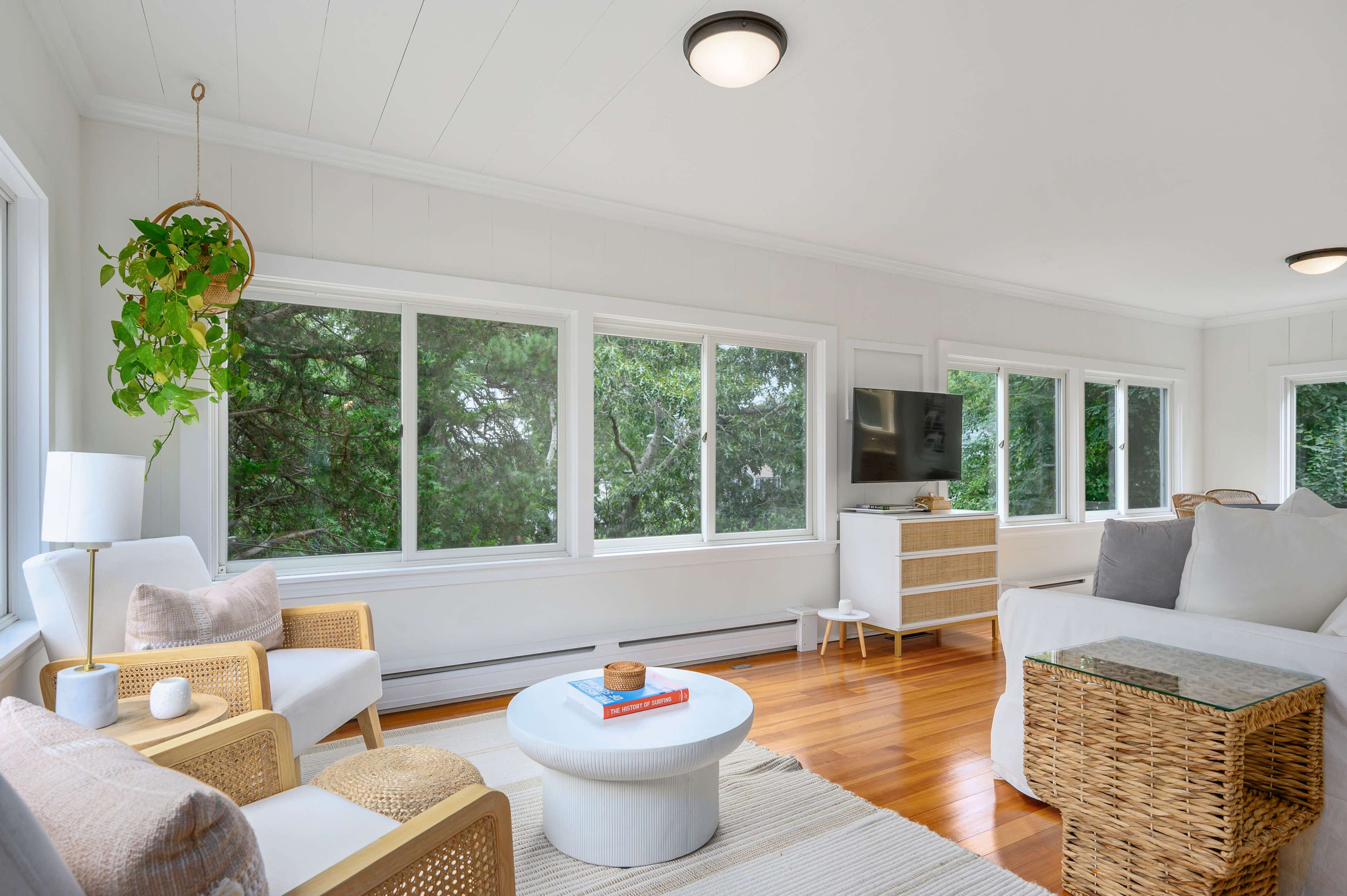 A bright living room features large windows, a light-colored sofa, woven furniture, and a small coffee table with a decorative item.