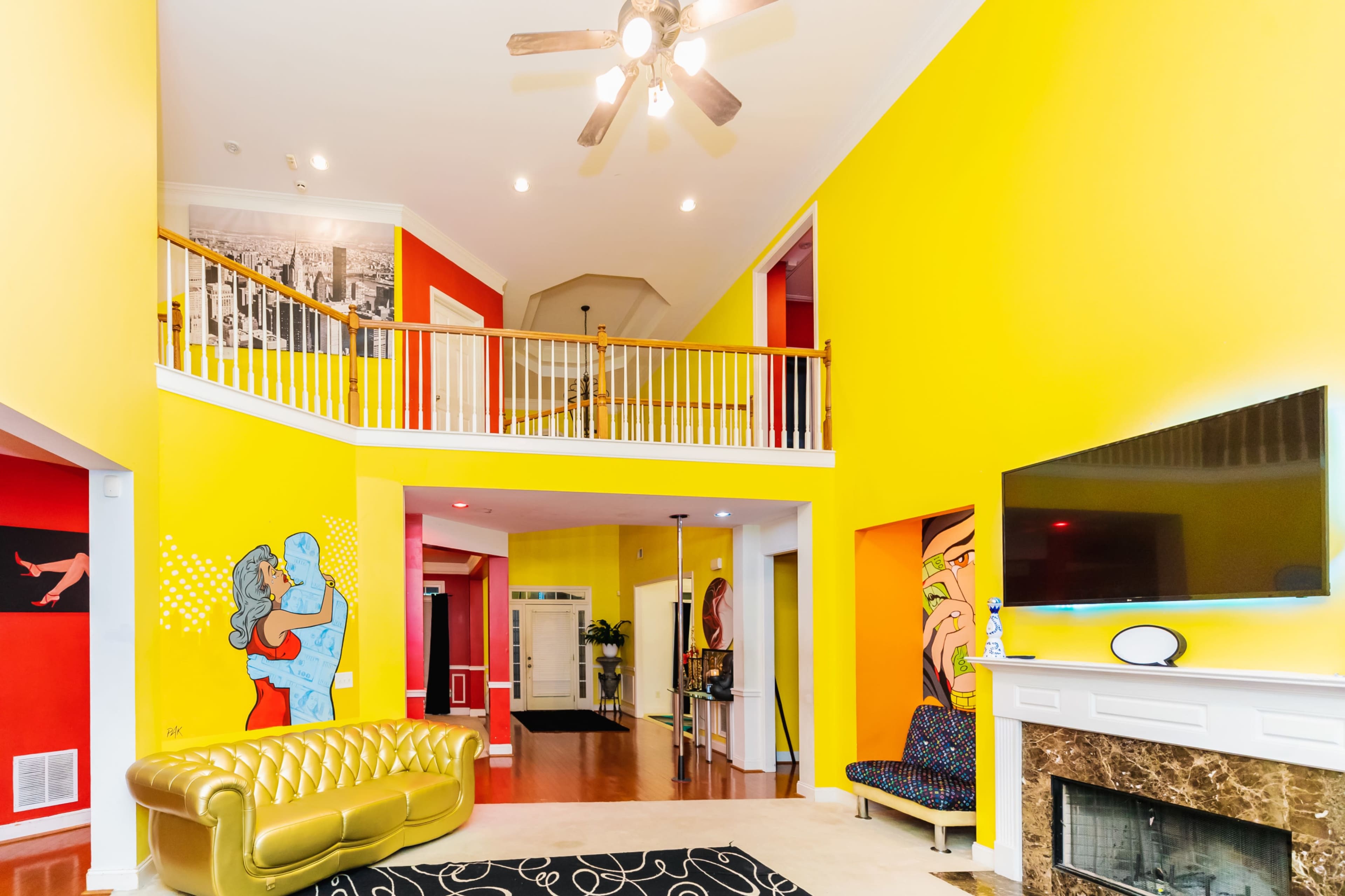 The interior features a spacious, brightly painted room with a high ceiling, a balcony, and colorful artwork on the walls.