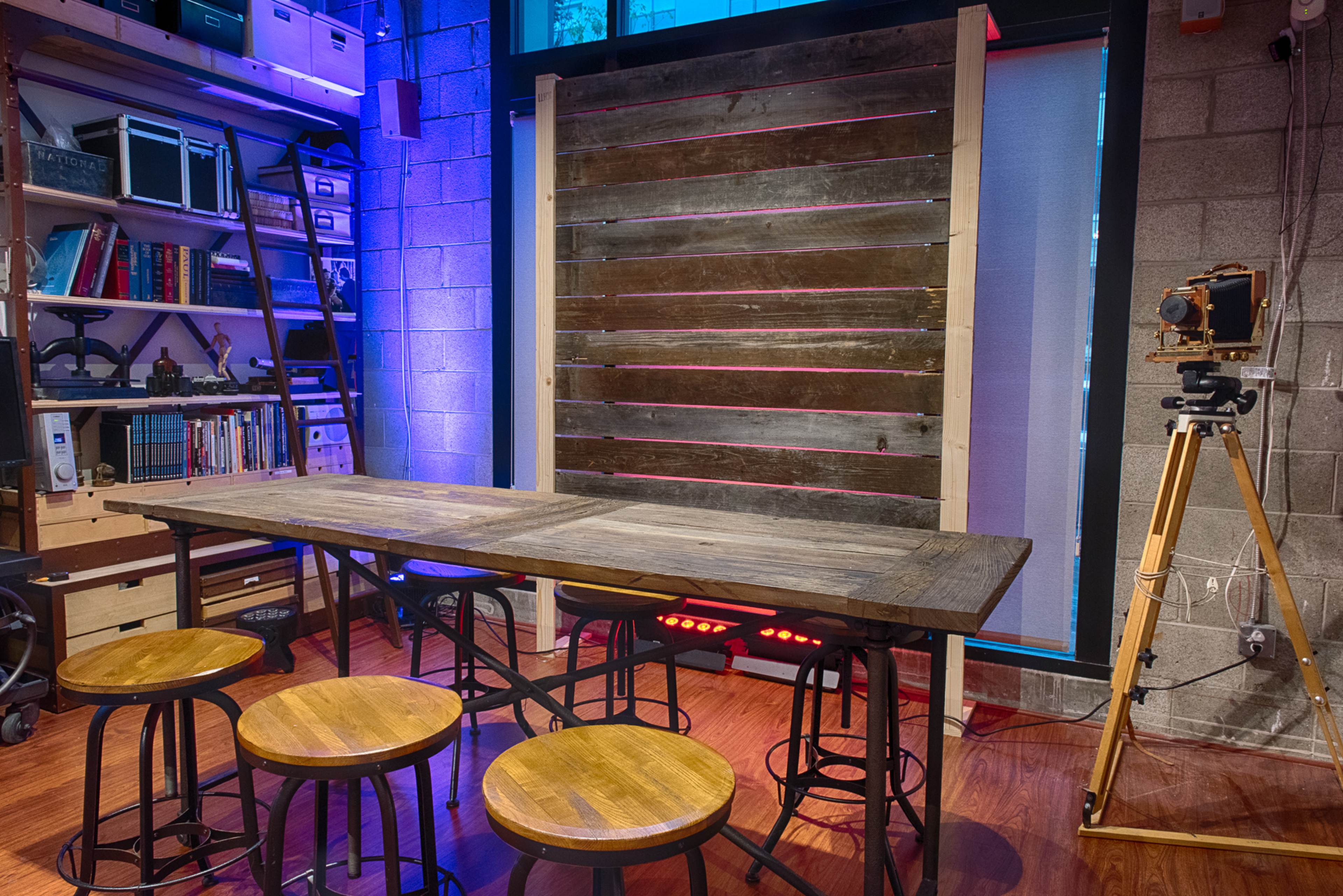 The scene features a wooden table surrounded by four stools, against a backdrop of a wooden wall with colored lighting and a vintage camera on a tripod.