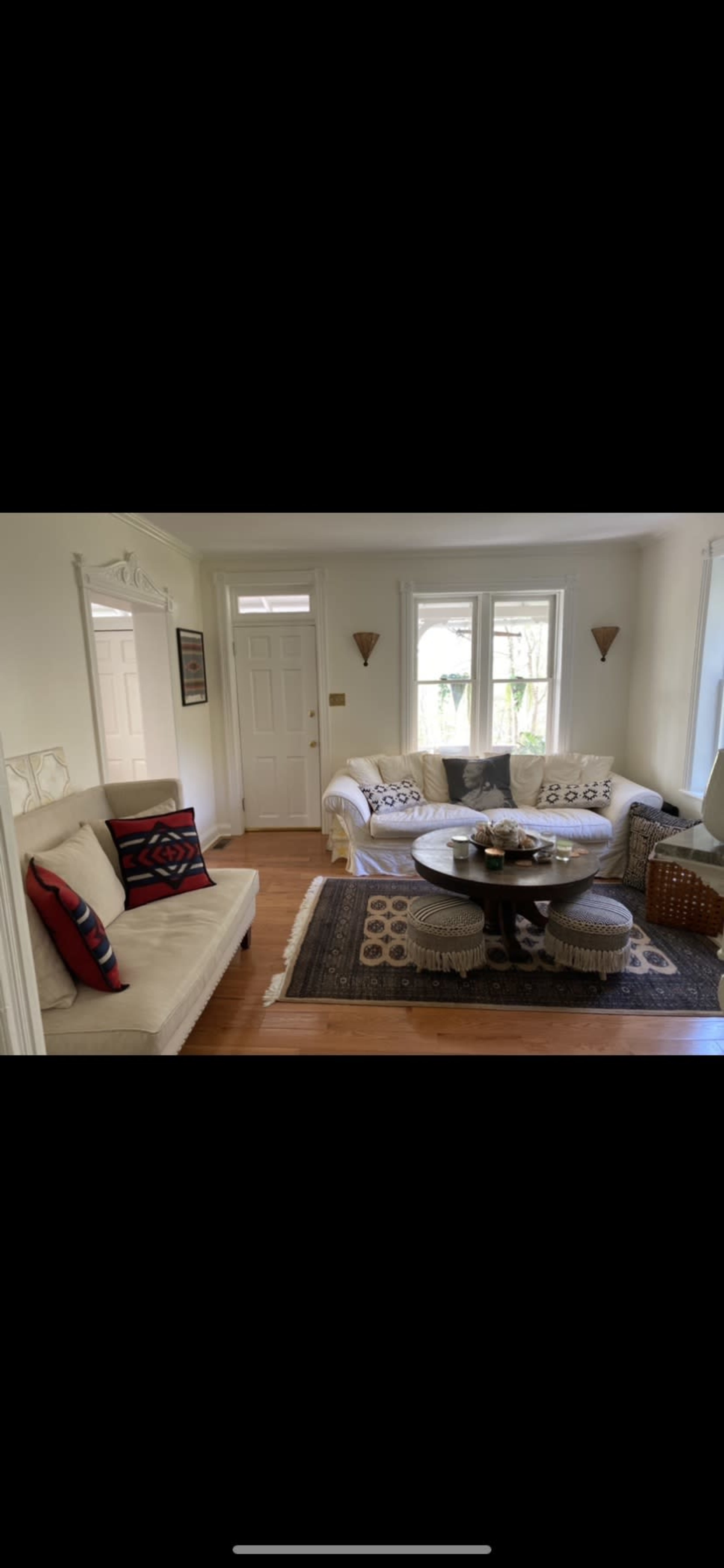 The image shows a living room with a white sofa, a circular coffee table, and a patterned rug, illuminated by natural light from the windows.