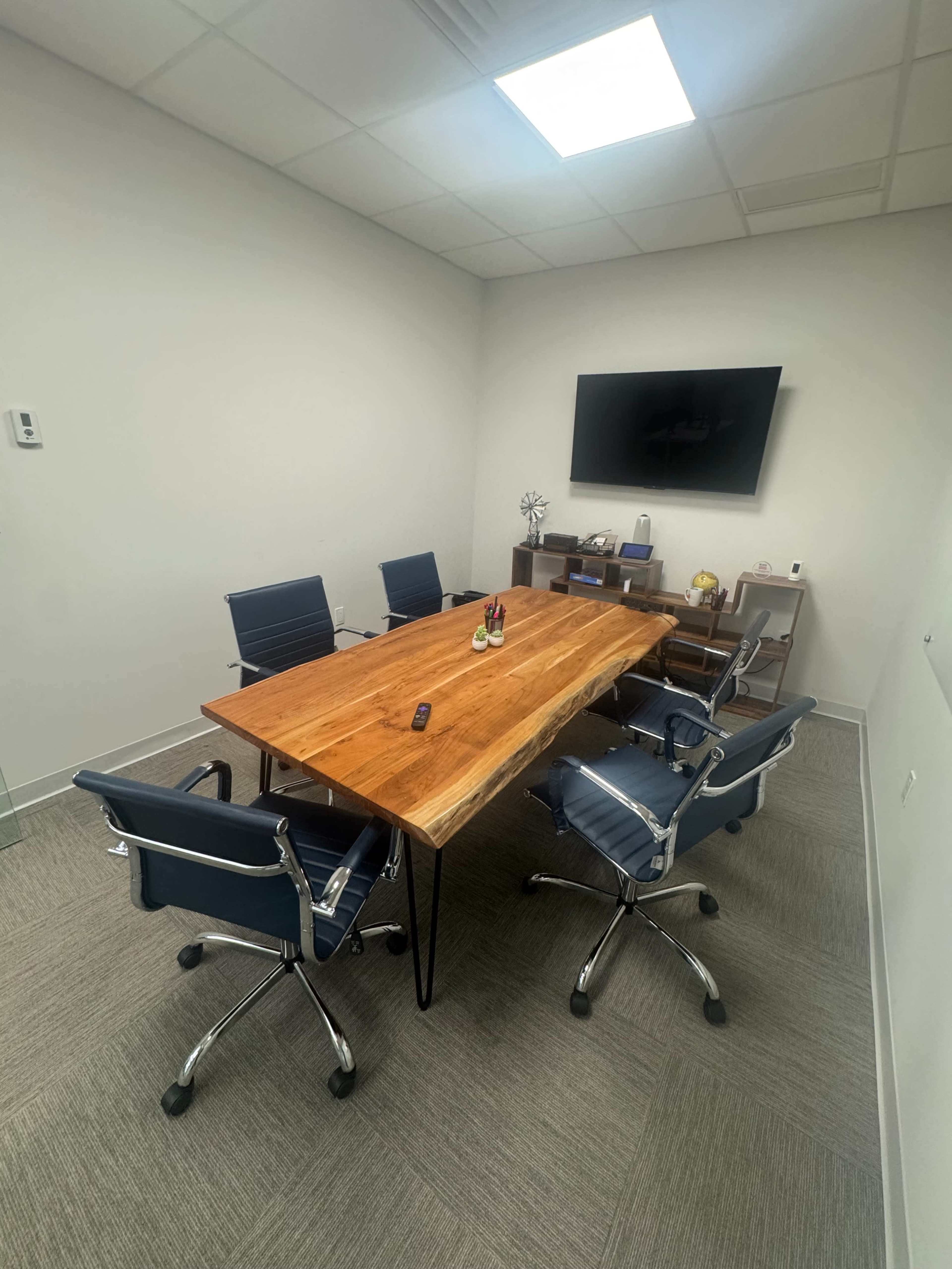 A conference room features a large wooden table surrounded by four blue office chairs and a television mounted on the wall.