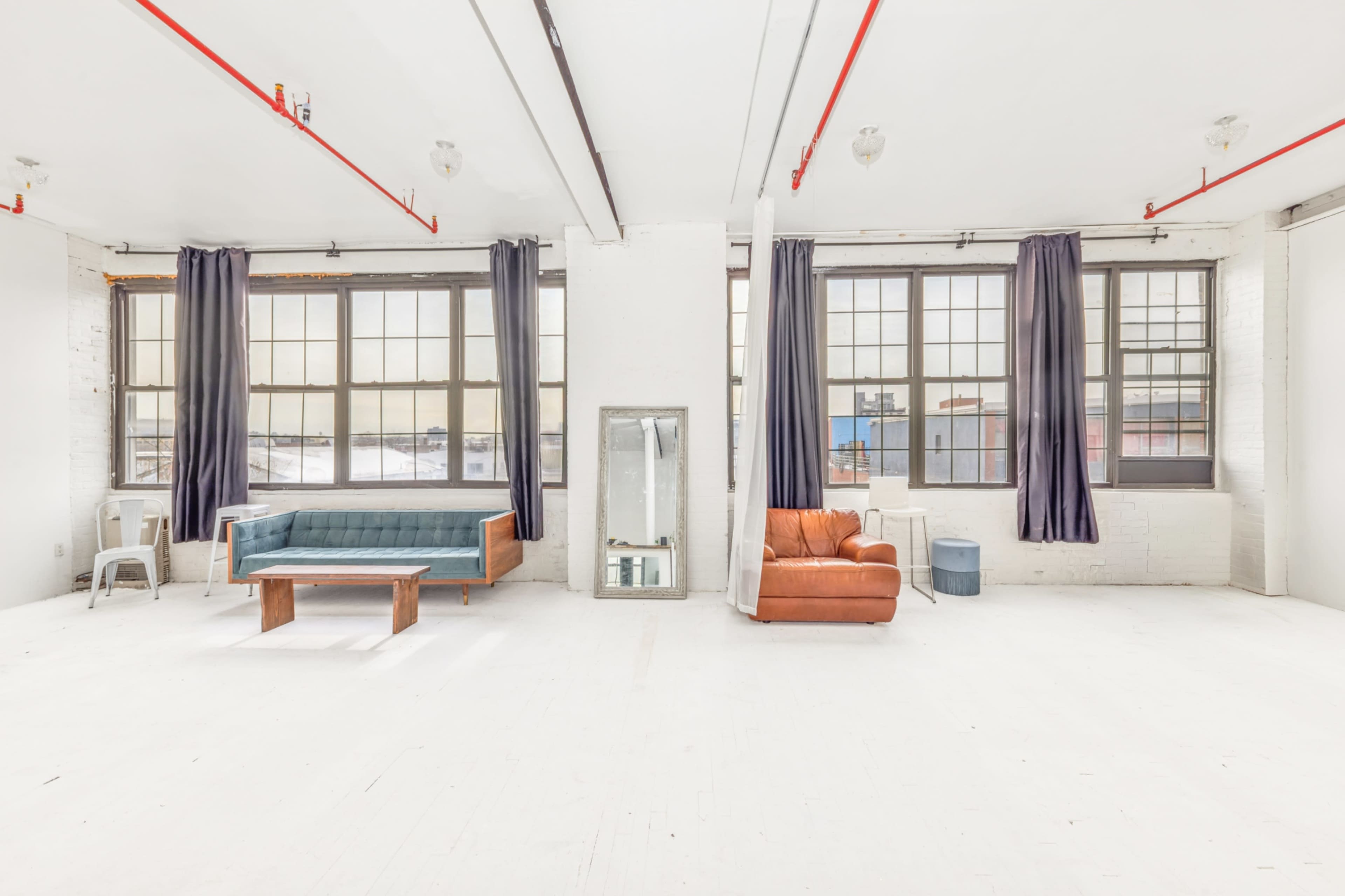 The image shows a bright, spacious loft with large windows, featuring a blue sofa, an orange armchair, a wooden coffee table, and a mirror.