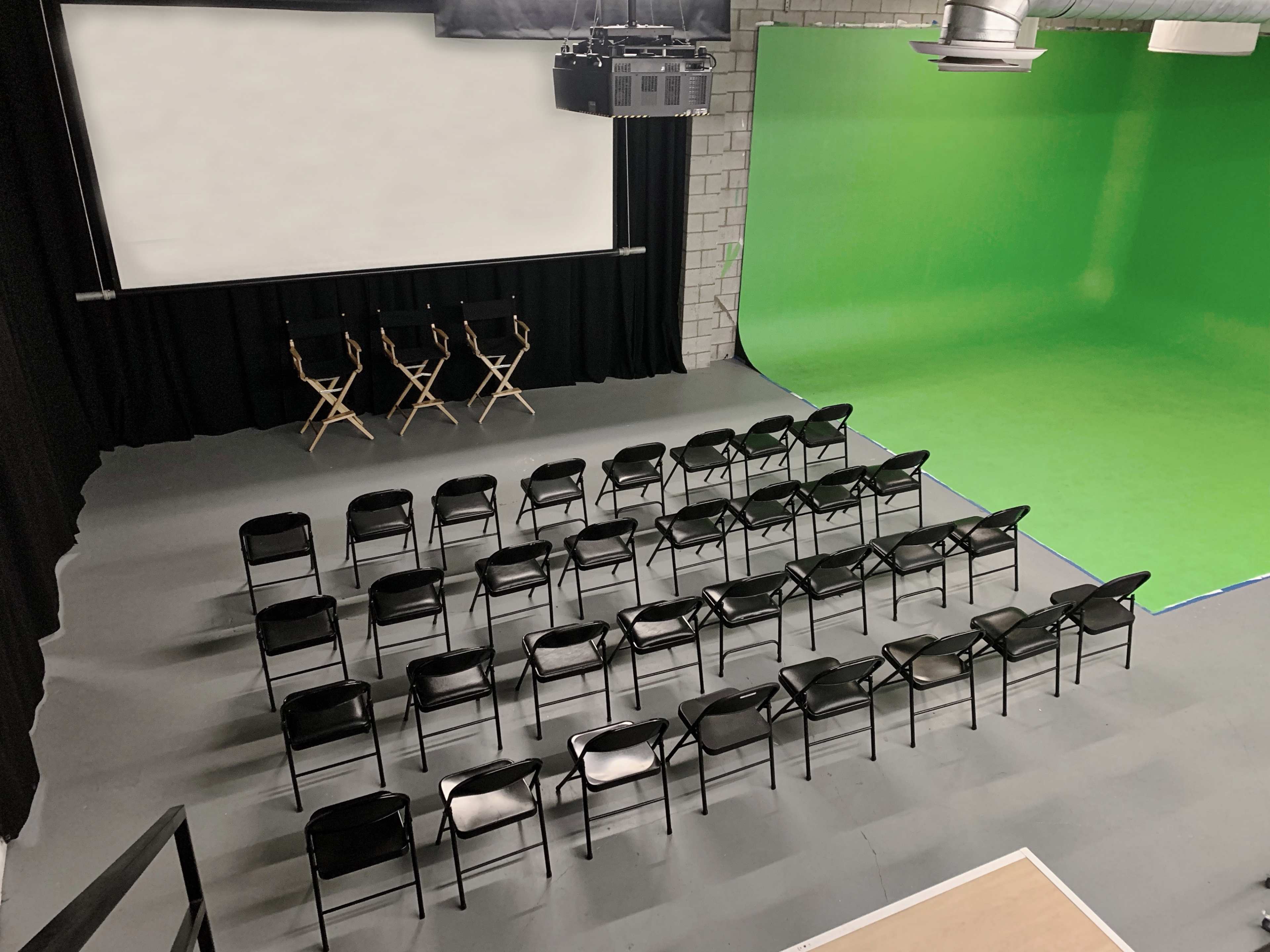 A studio setup with a green screen on one wall, a blank projection screen on another, and rows of black chairs arranged for an audience.