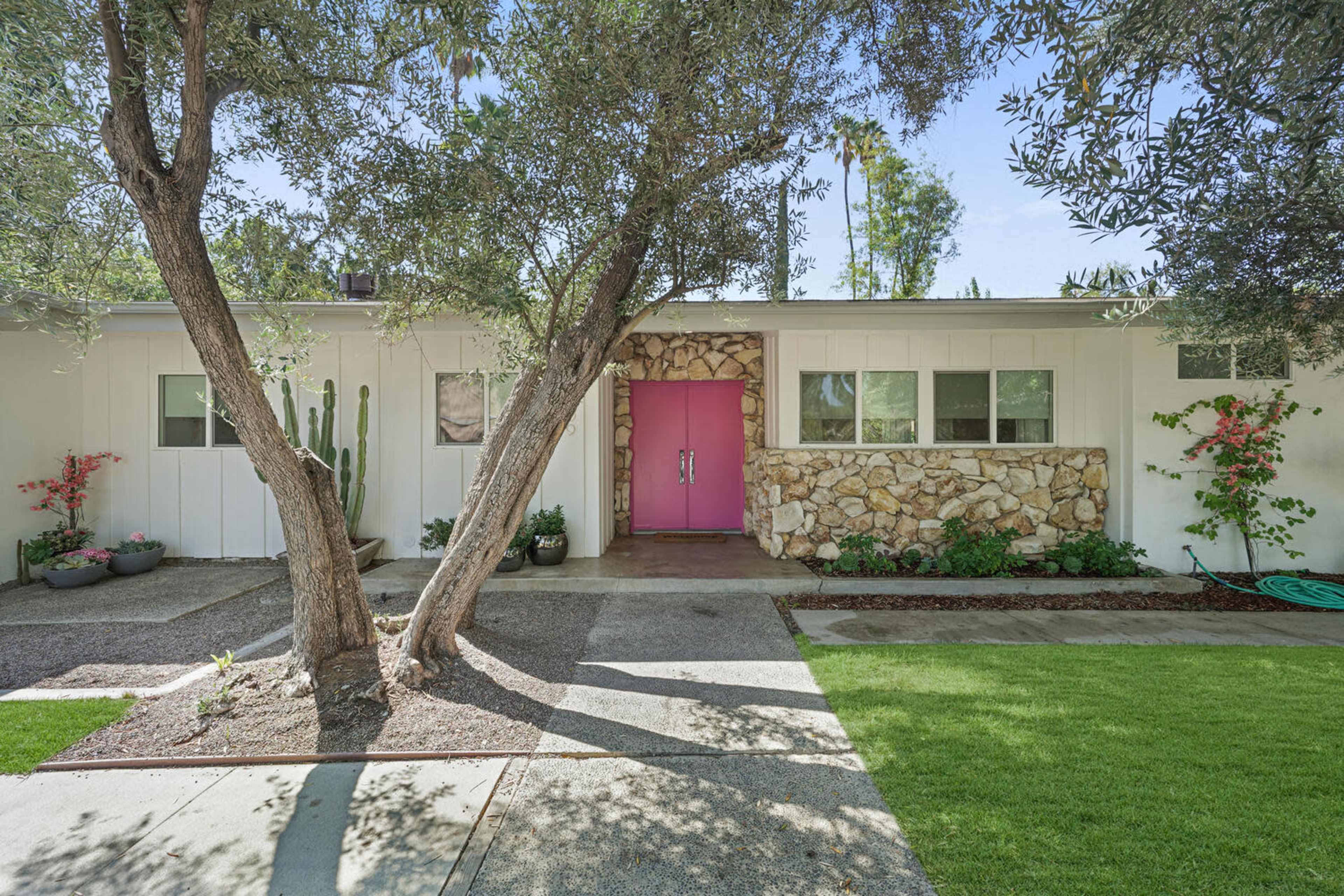 A modern home features a stone accent wall and a vibrant pink front door, framed by lush greenery and landscaping.