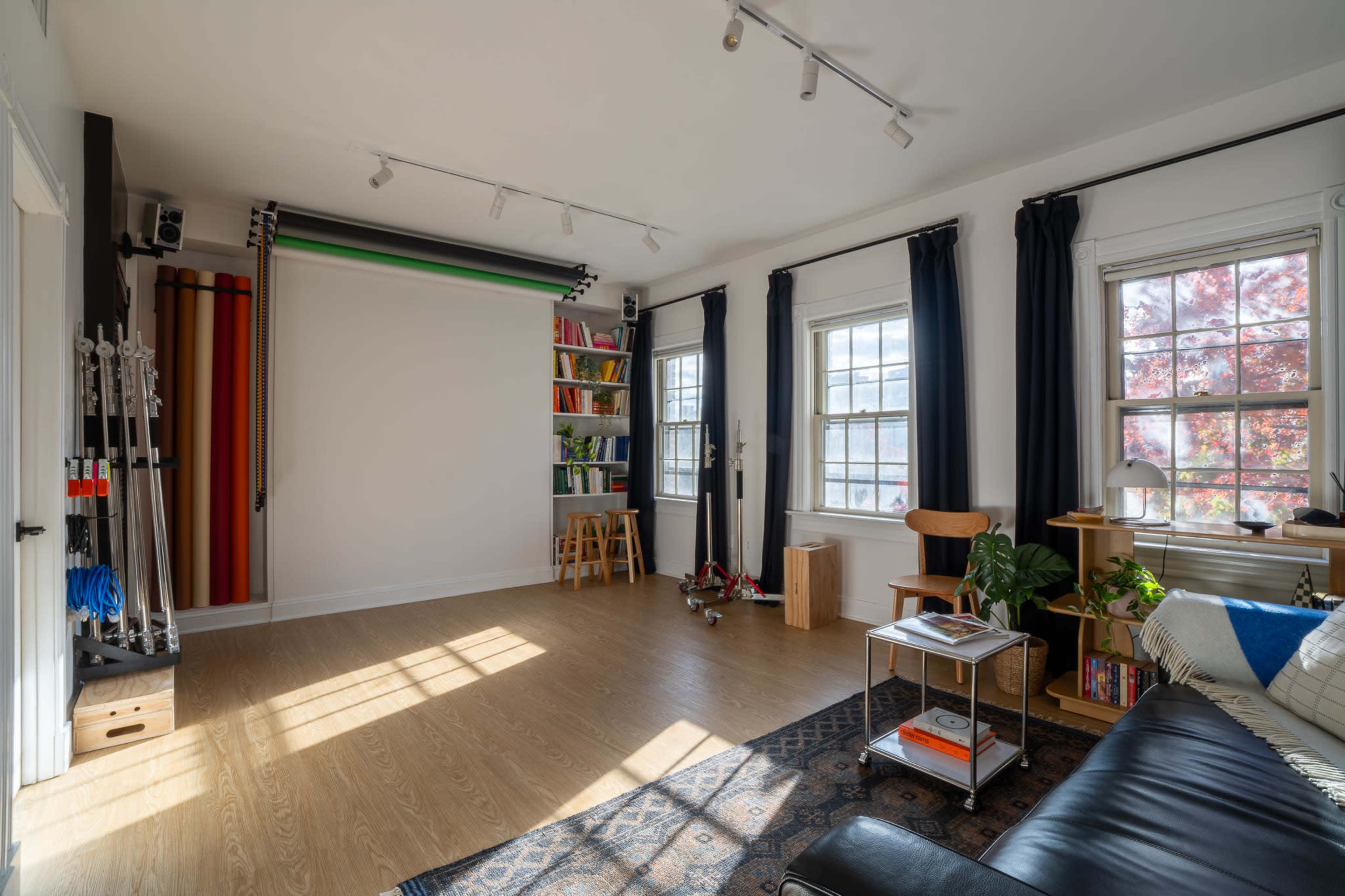 The room features a hardwood floor, large windows with curtains, a bookshelf filled with colorful books, and a backdrop setup for photography.