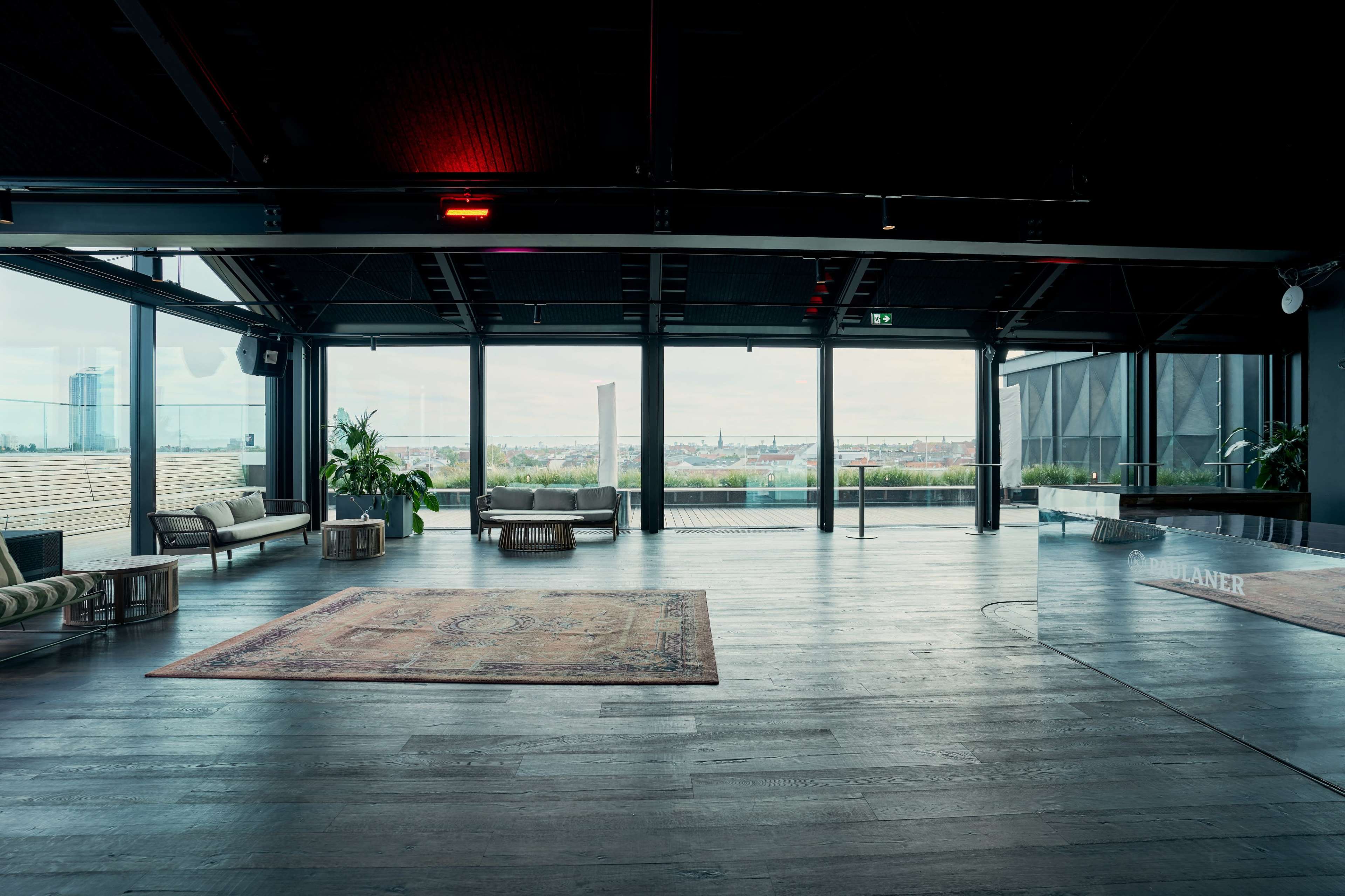 The image shows a spacious, modern interior with wooden floors, large windows offering a view of the city, and a few pieces of furniture, including a rug and sofas.