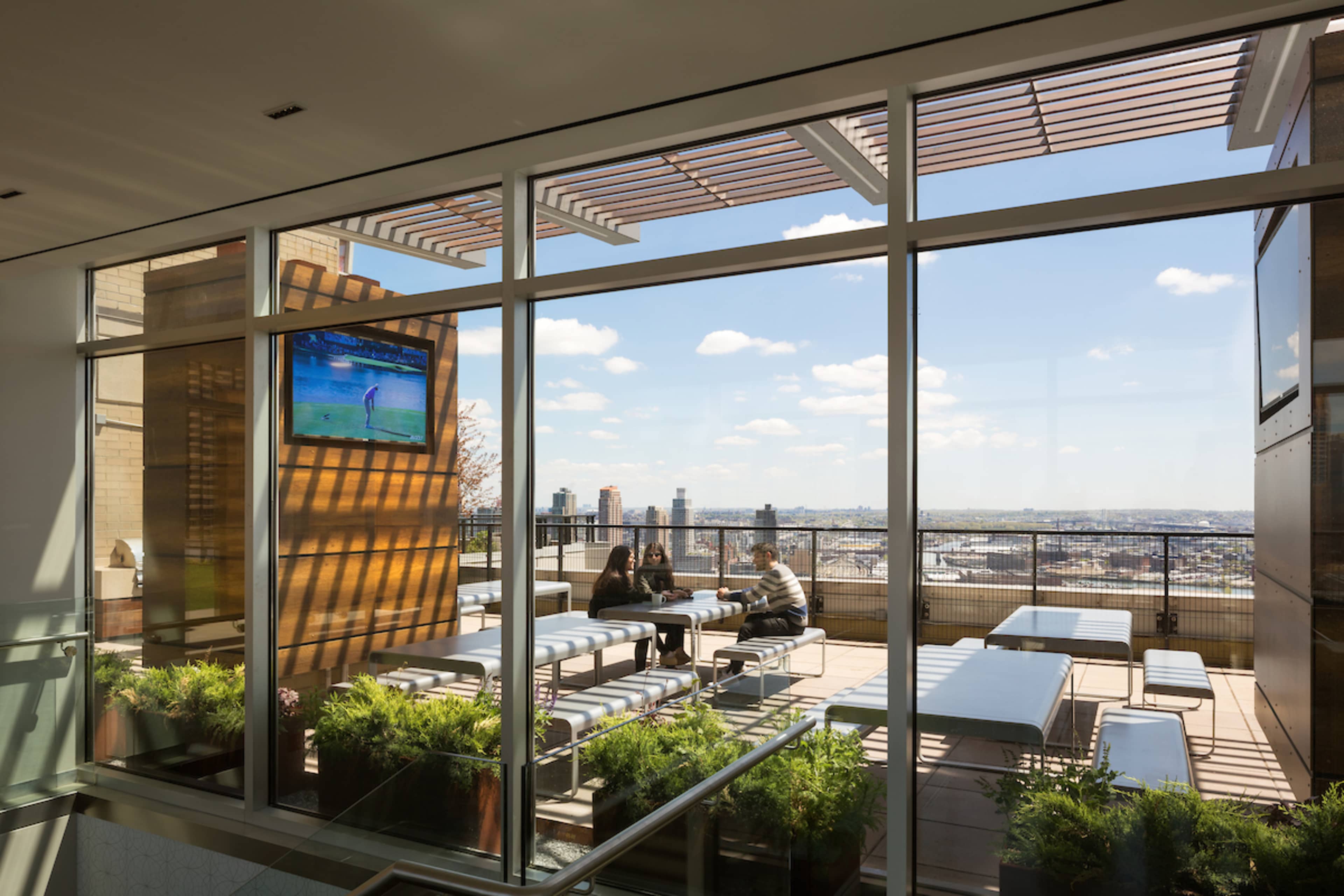 A modern rooftop terrace with outdoor seating and a television, featuring a view of the city skyline through large glass windows.