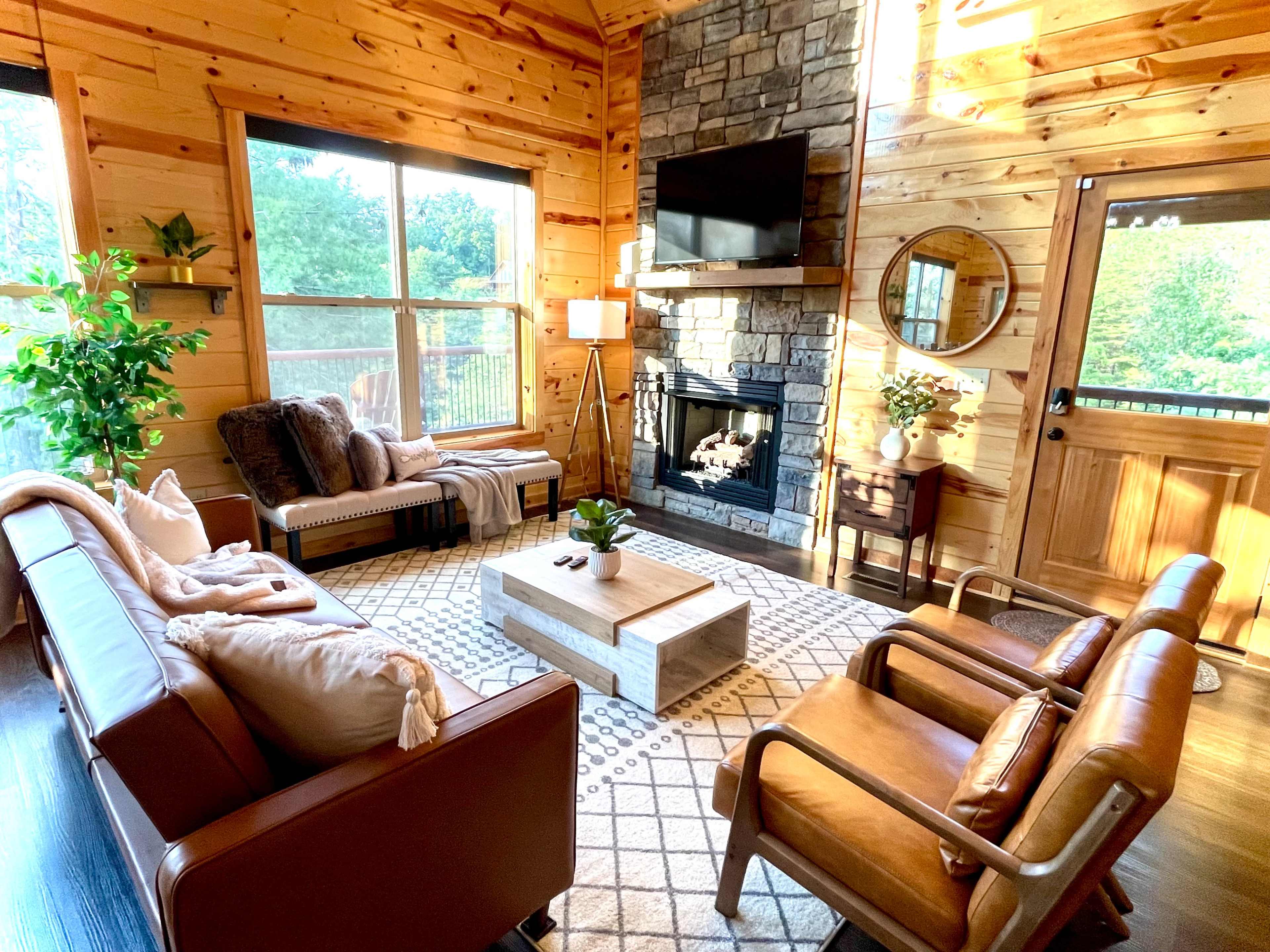 The image shows a cozy living room with natural wood walls, a stone fireplace, a coffee table in the center, and two comfortable seating areas arranged around a rug.