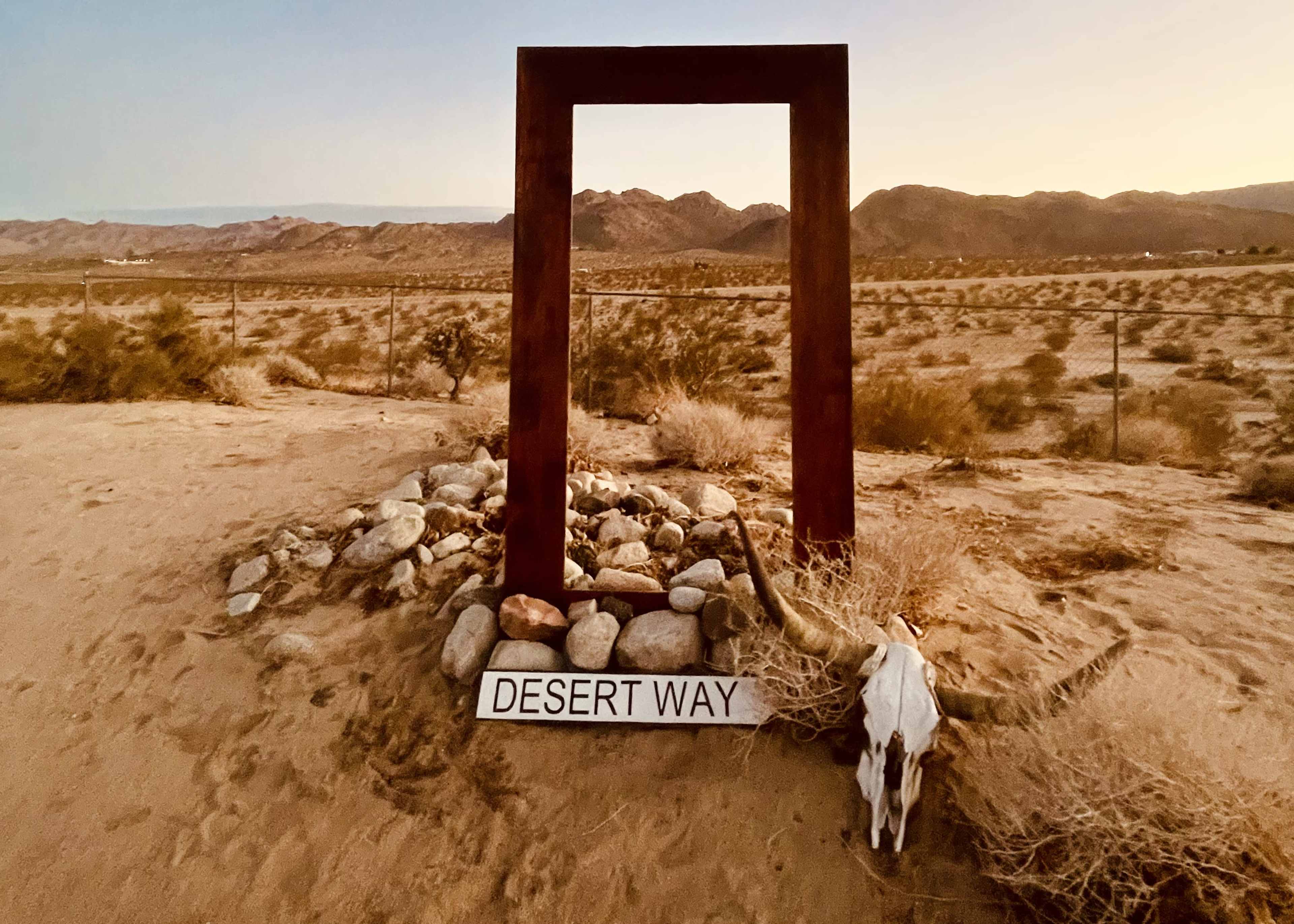 A large rectangular frame sits on the desert ground, surrounded by rocks, with a sign reading "DESERT WAY" and a skull nearby, set against a backdrop of mountains.