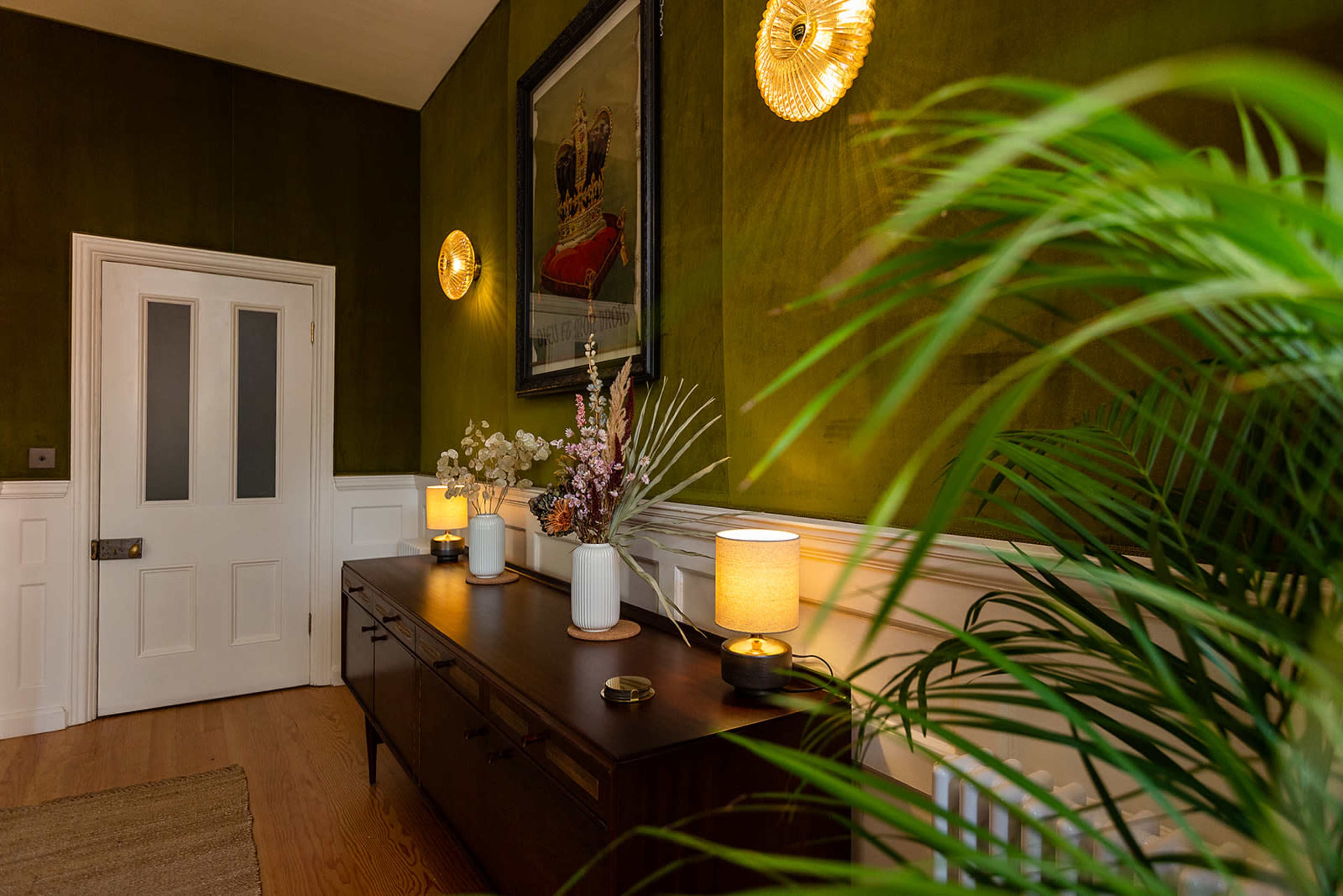 A hallway features a dark green wall with a sideboard, decorative lamps, a framed artwork, and a potted plant.