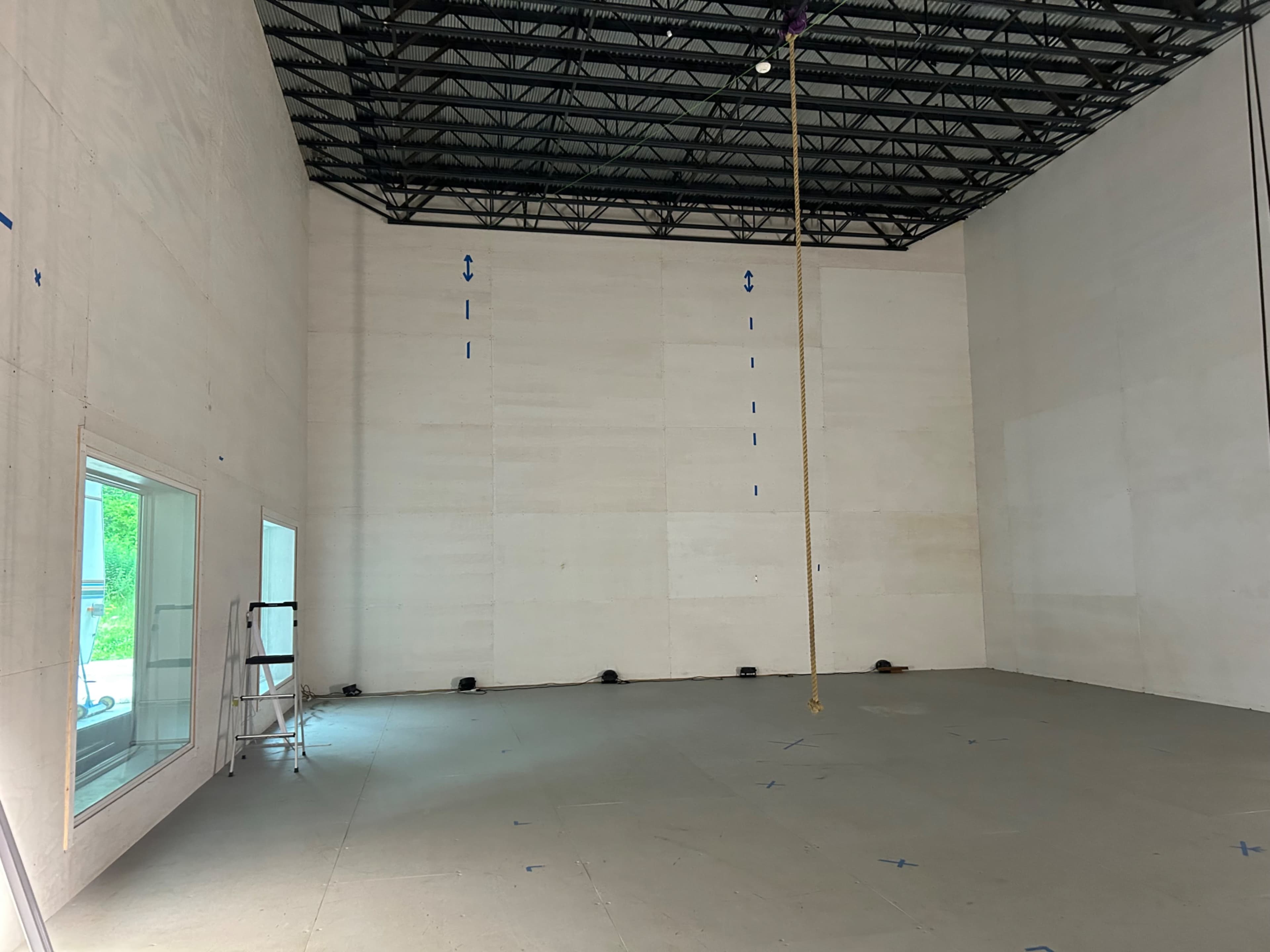 An empty, high-ceilinged interior space with bare walls and a tall rope hanging from the ceiling.