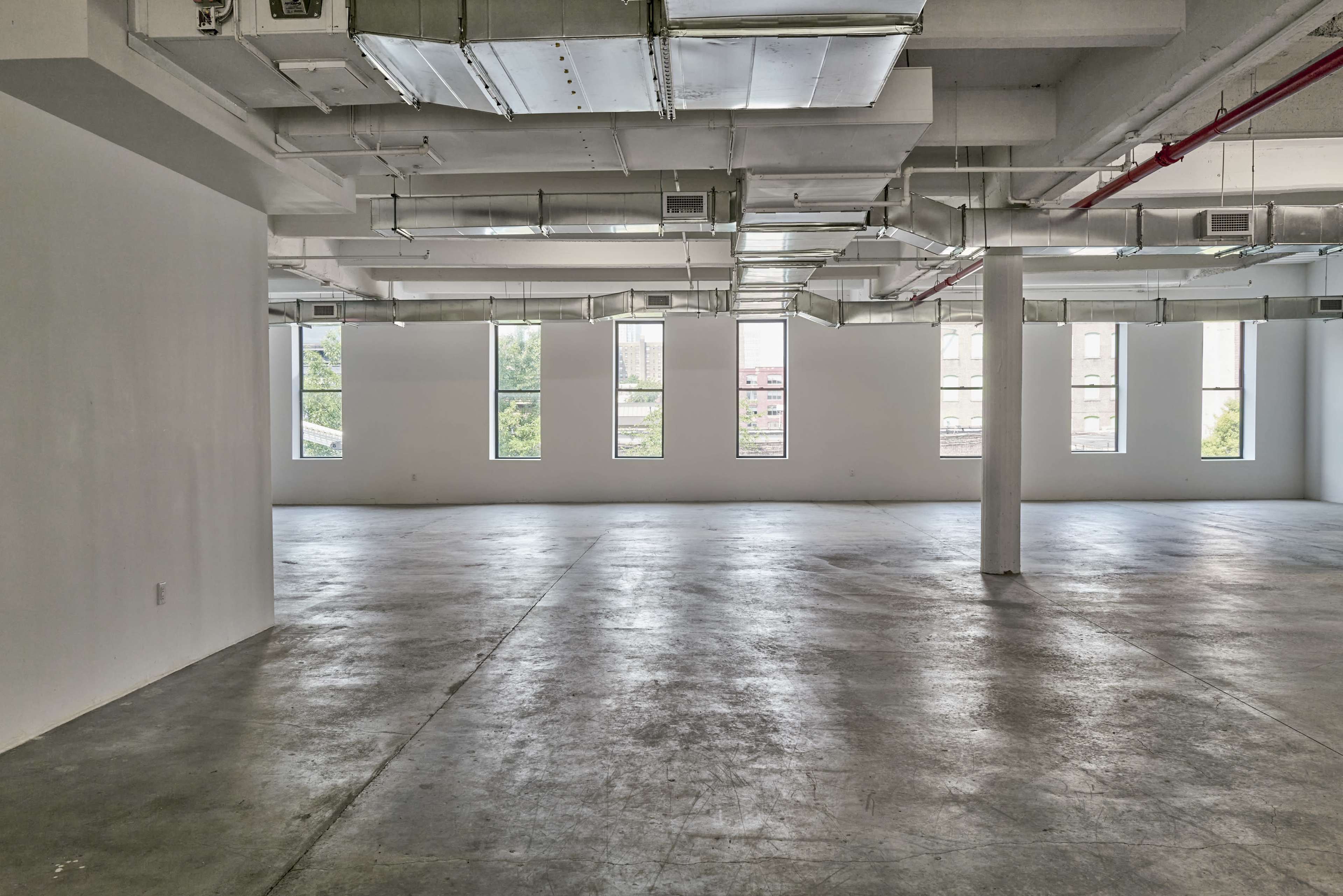 The image shows a spacious, empty room with large windows lined along the walls and concrete flooring.