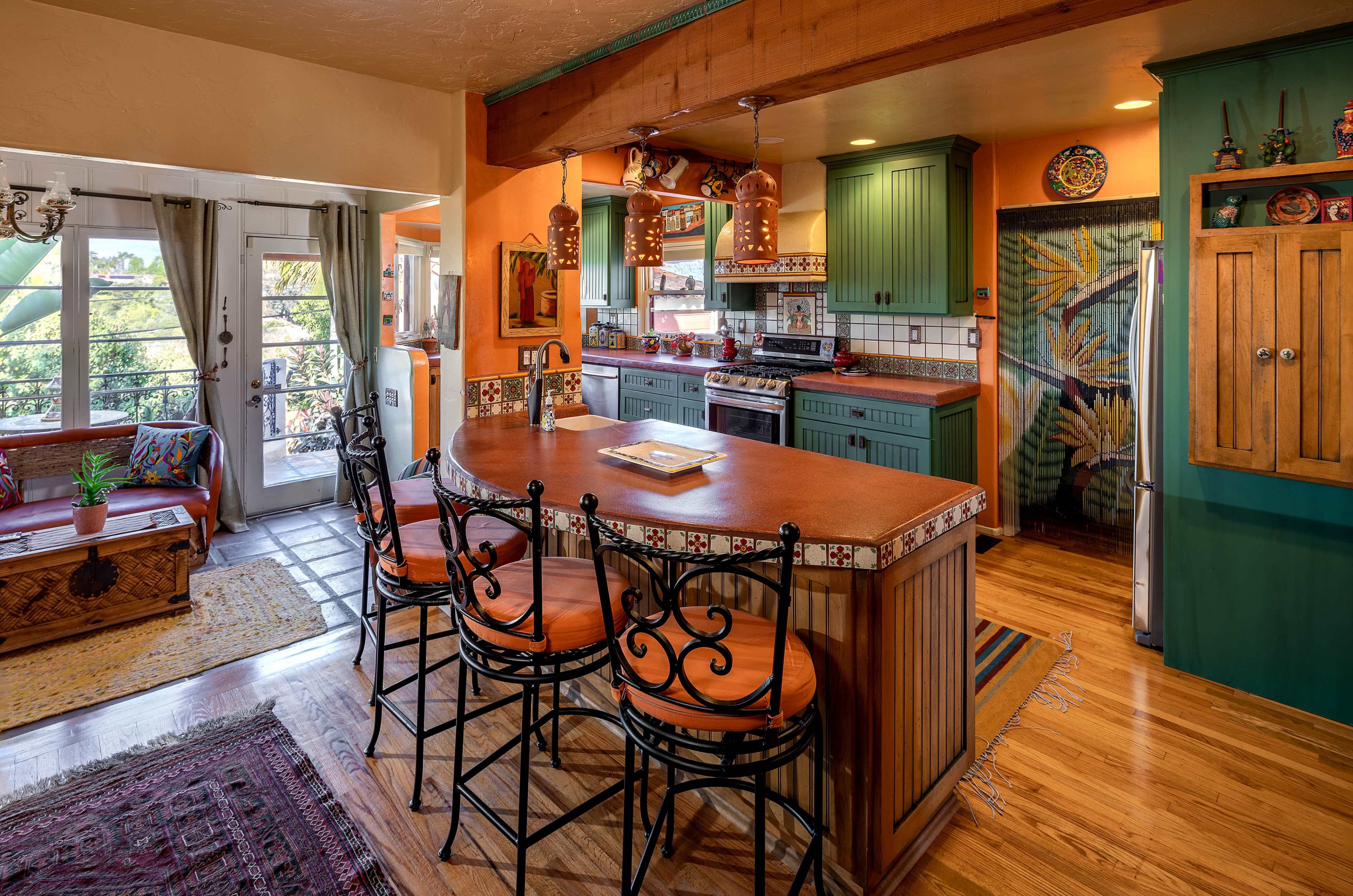 A colorful kitchen with wooden cabinetry, a tiled countertop, and a breakfast bar with high-backed chairs, overlooking a living area with a sofa and decorative accents.