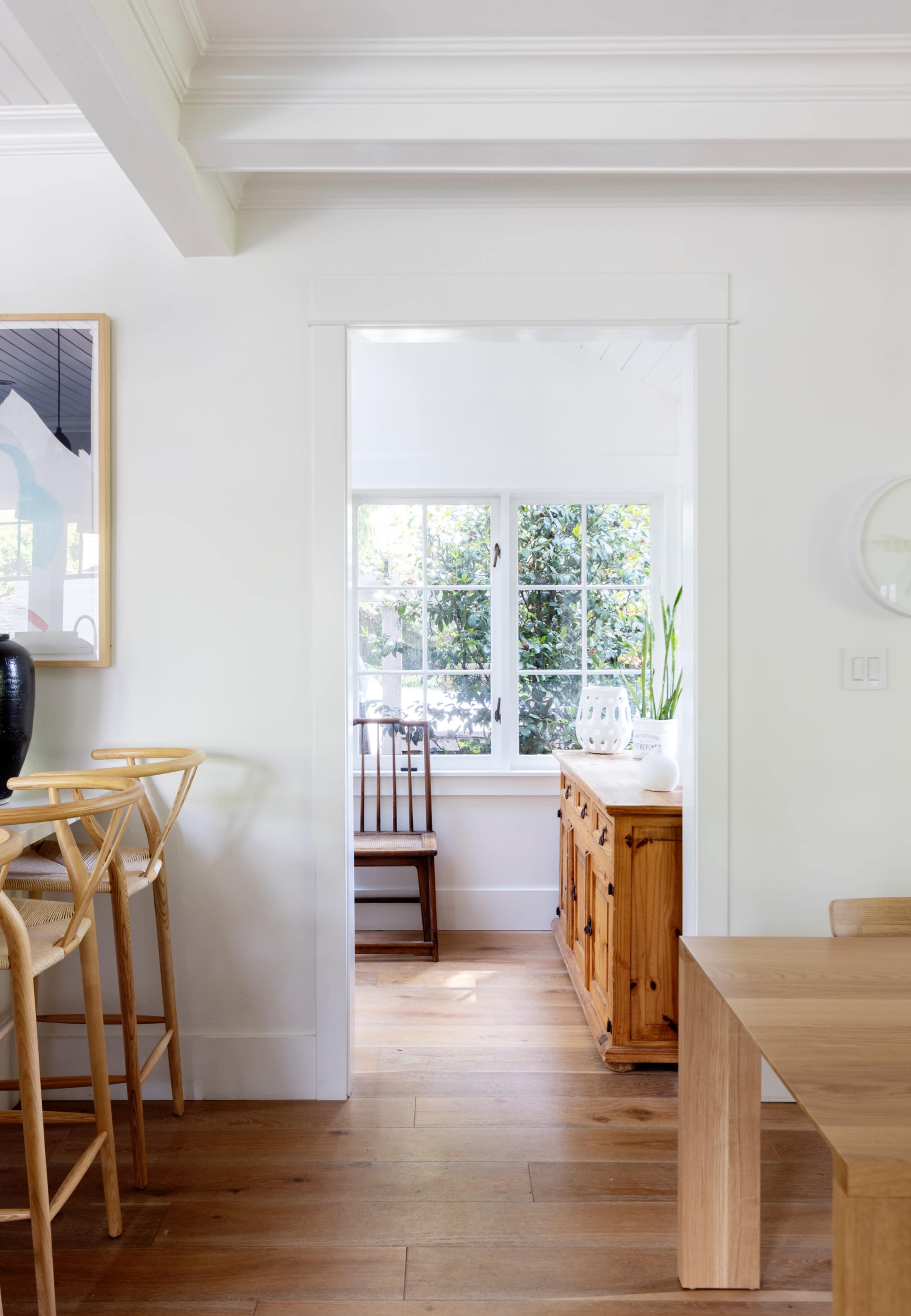 A bright, airy interior space with a wooden side table, a tall chair, and a view into a sunlit room featuring a wooden cabinet and plants.