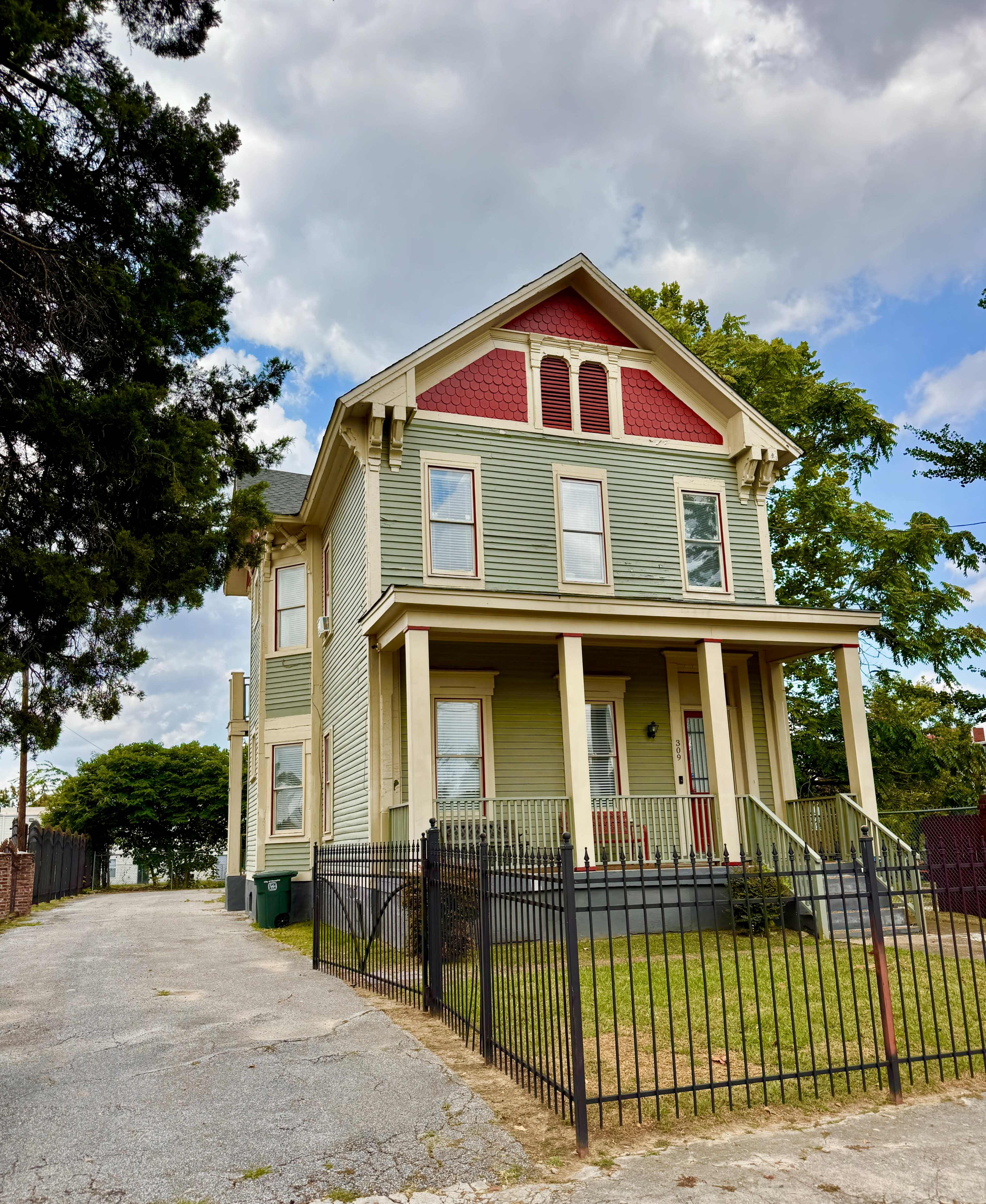 A large, two-story Victorian-style house features green and red siding, a manicured lawn, and a gated porch.