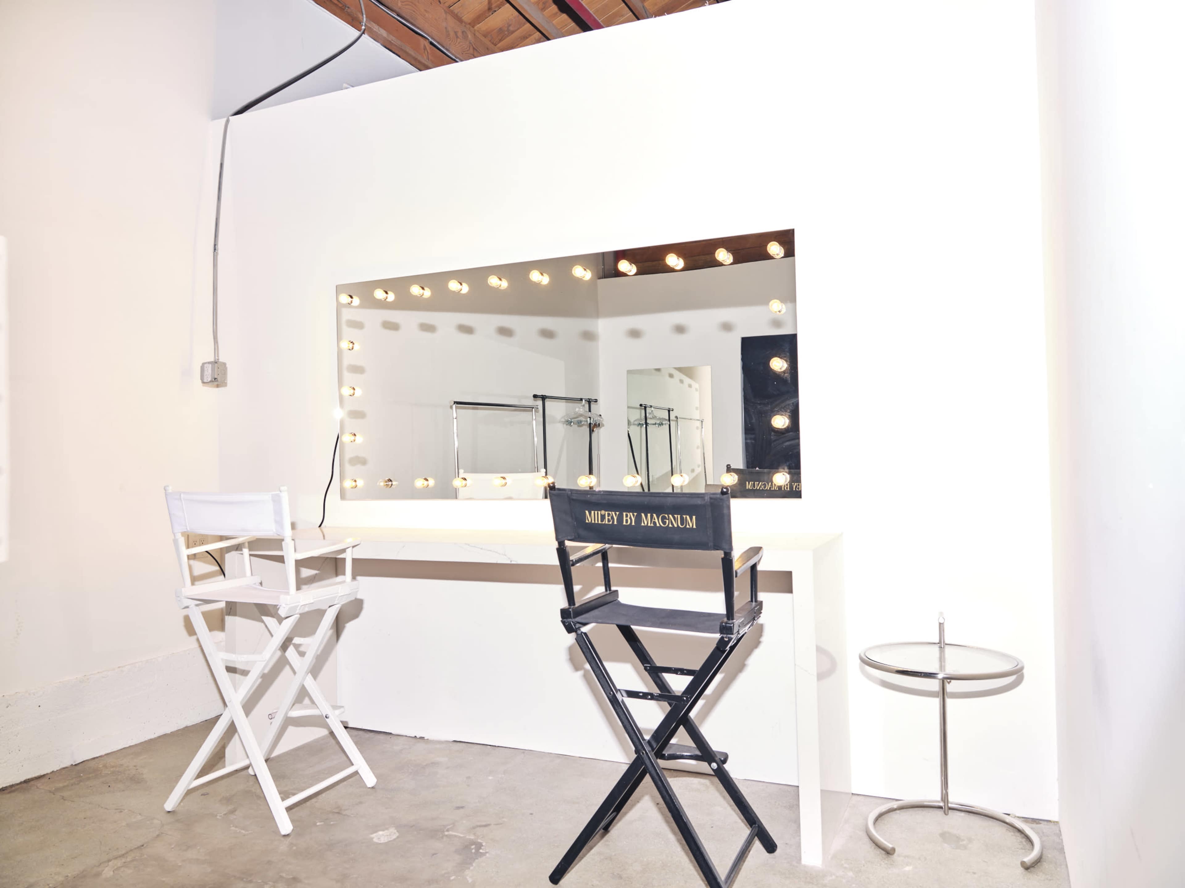 The image shows a minimalist makeup vanity with a large illuminated mirror, two director's chairs, and a small table in a well-lit room.