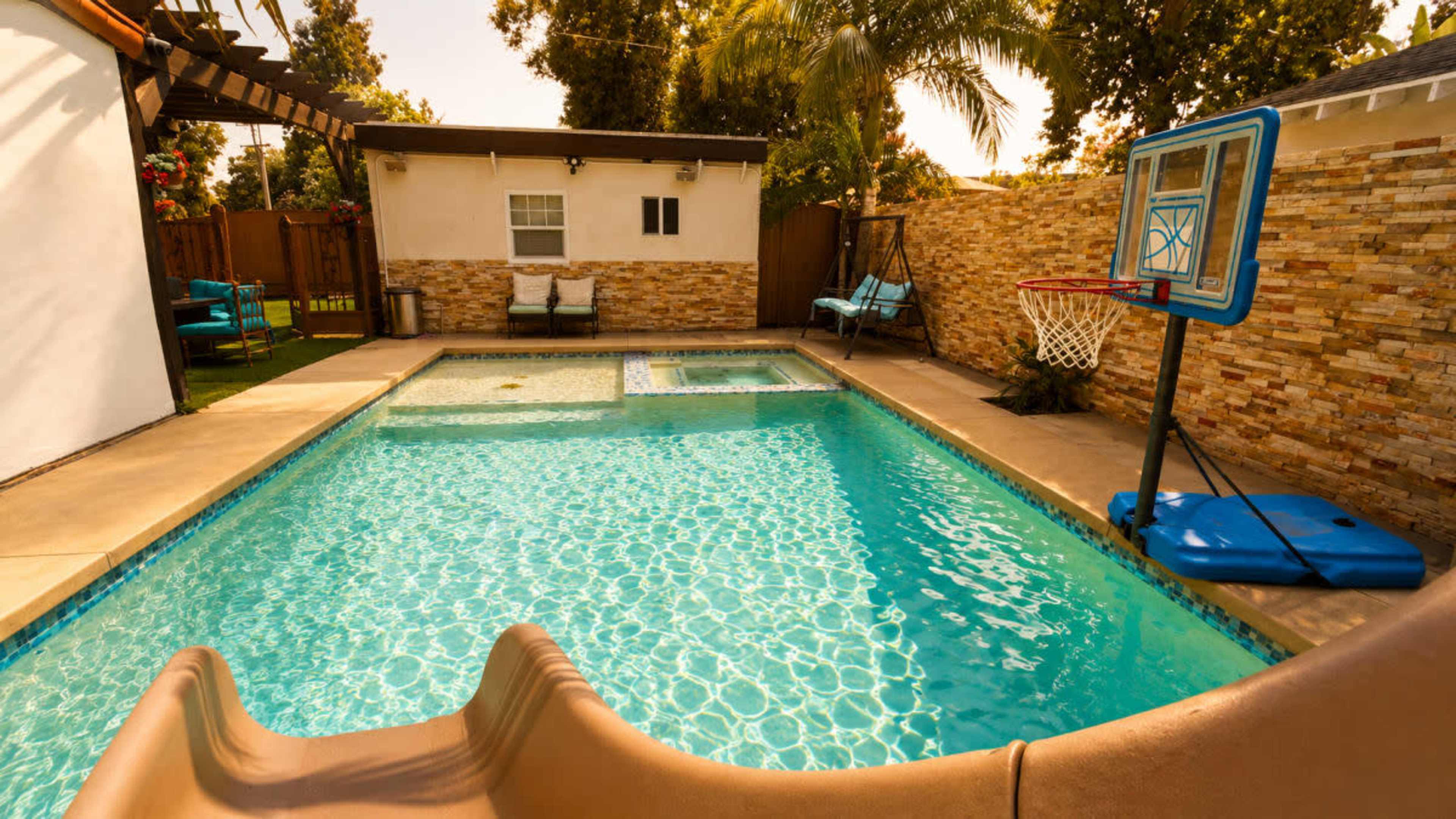The image shows a swimming pool with a slide, a spa area, and a basketball hoop, surrounded by landscaped greenery and a stone wall.