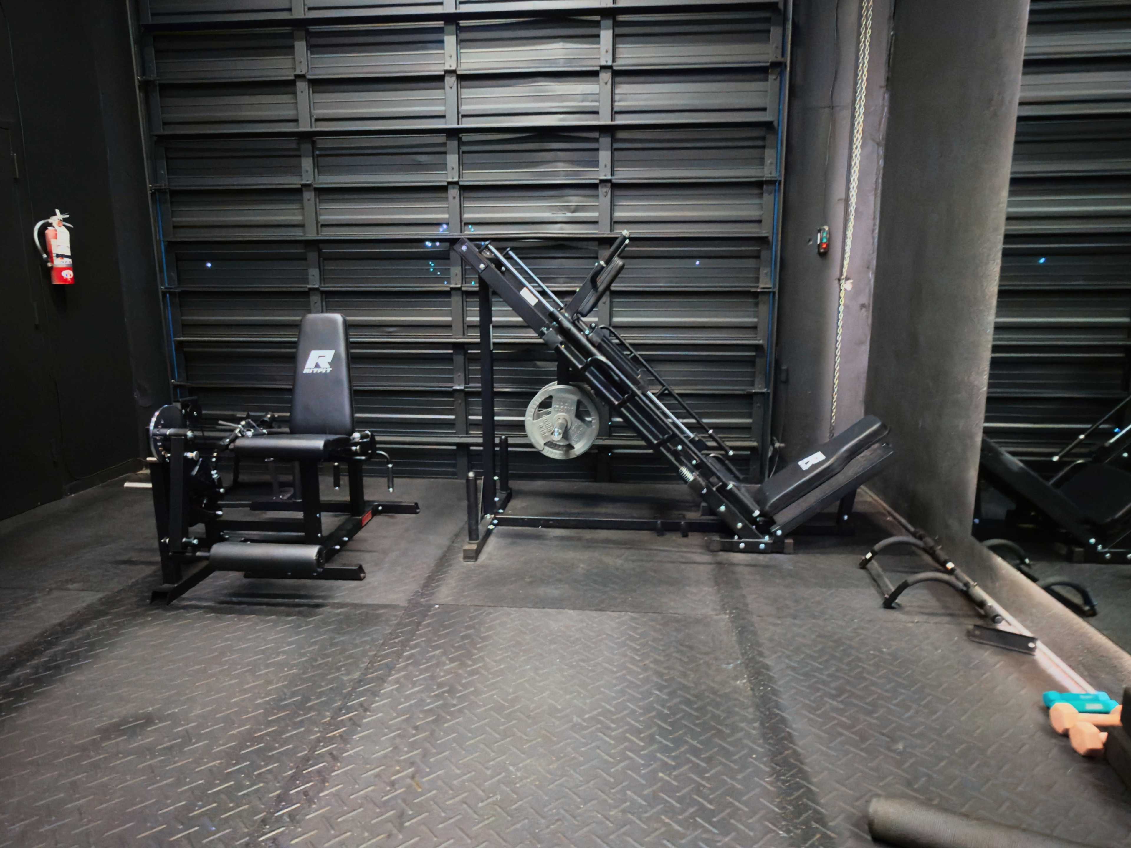 The image shows a gym interior featuring a leg press machine on the right and a seated leg curl machine on the left, against a dark metal backdrop.